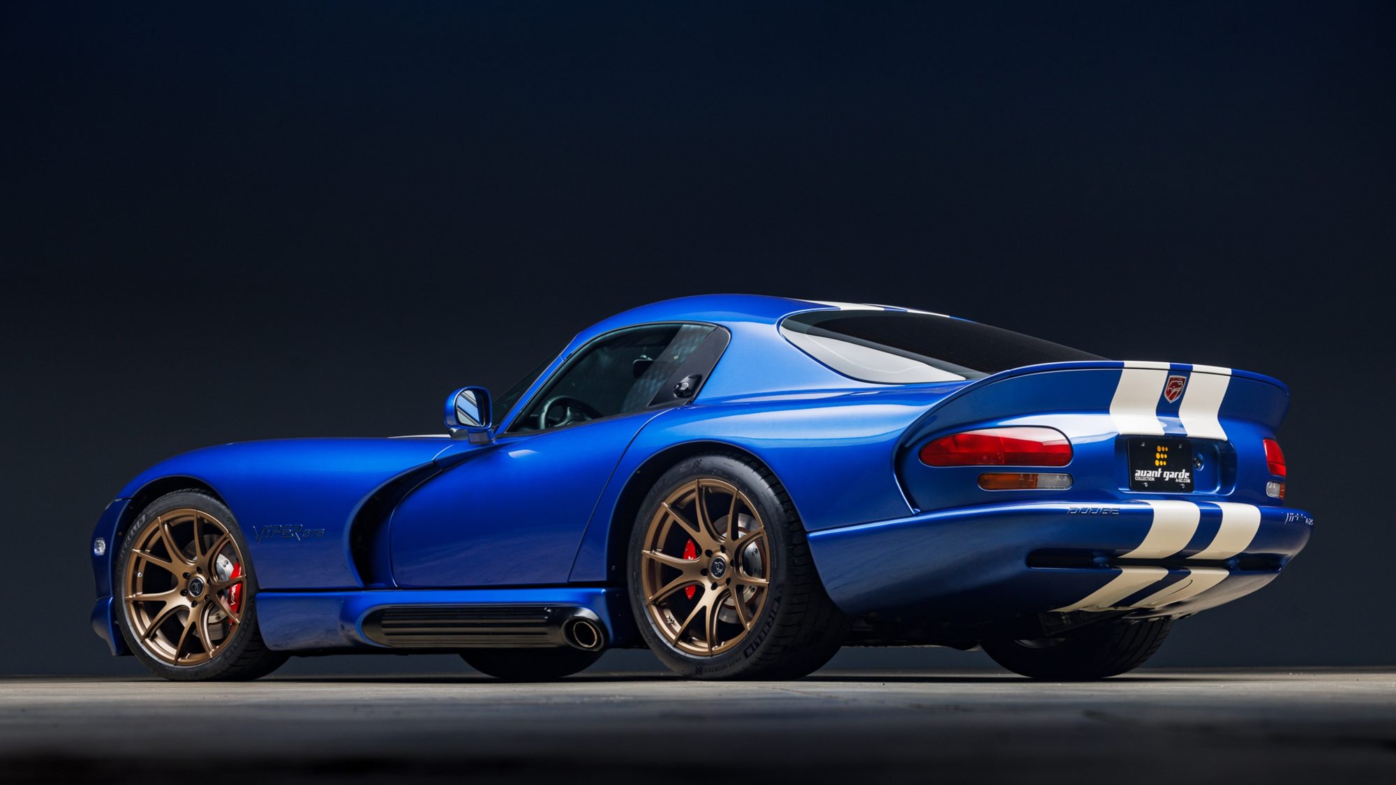 A blue Dodge Viper GTS coupe seen from the rear three-quarter angle in a studio, showing its wide rear fenders, side-exit exhaust, bronze wheels and white stripes over the tail.