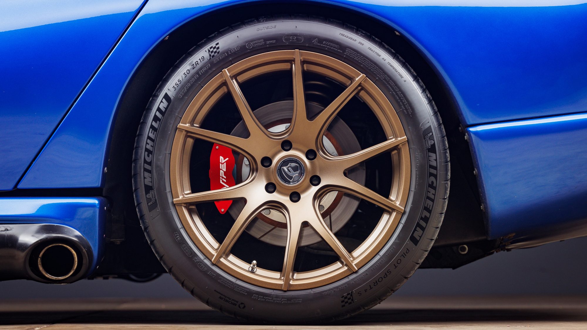 A close view of a bronze aftermarket wheel on a blue Dodge Viper, with a red brake caliper lettered Viper, a Michelin tire and the side-exit exhaust tip beside it.