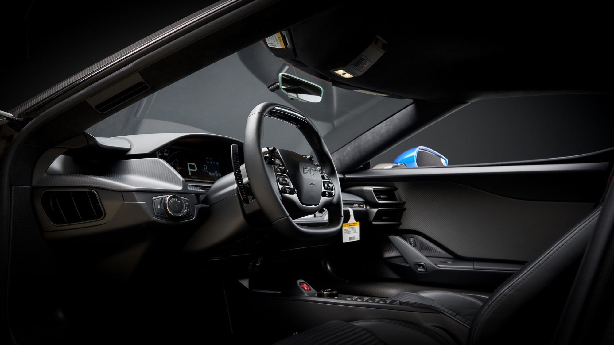 The Ford GT's dark cockpit seen through the open driver's door: a squared-off steering wheel with a GT badge and buttons, the digital gauge cluster showing P, and carbon fiber trim