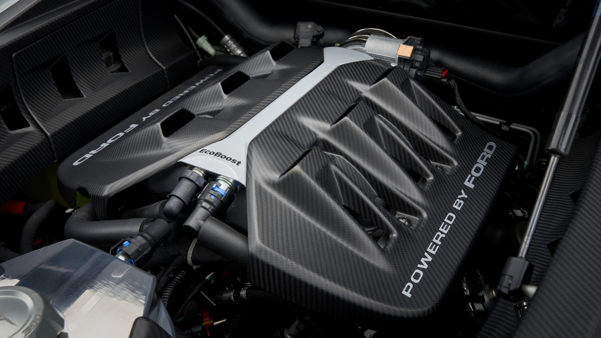 Close-up of the Ford GT's matte carbon fiber engine covers, marked POWERED BY FORD and EcoBoost, with hoses and connectors around them