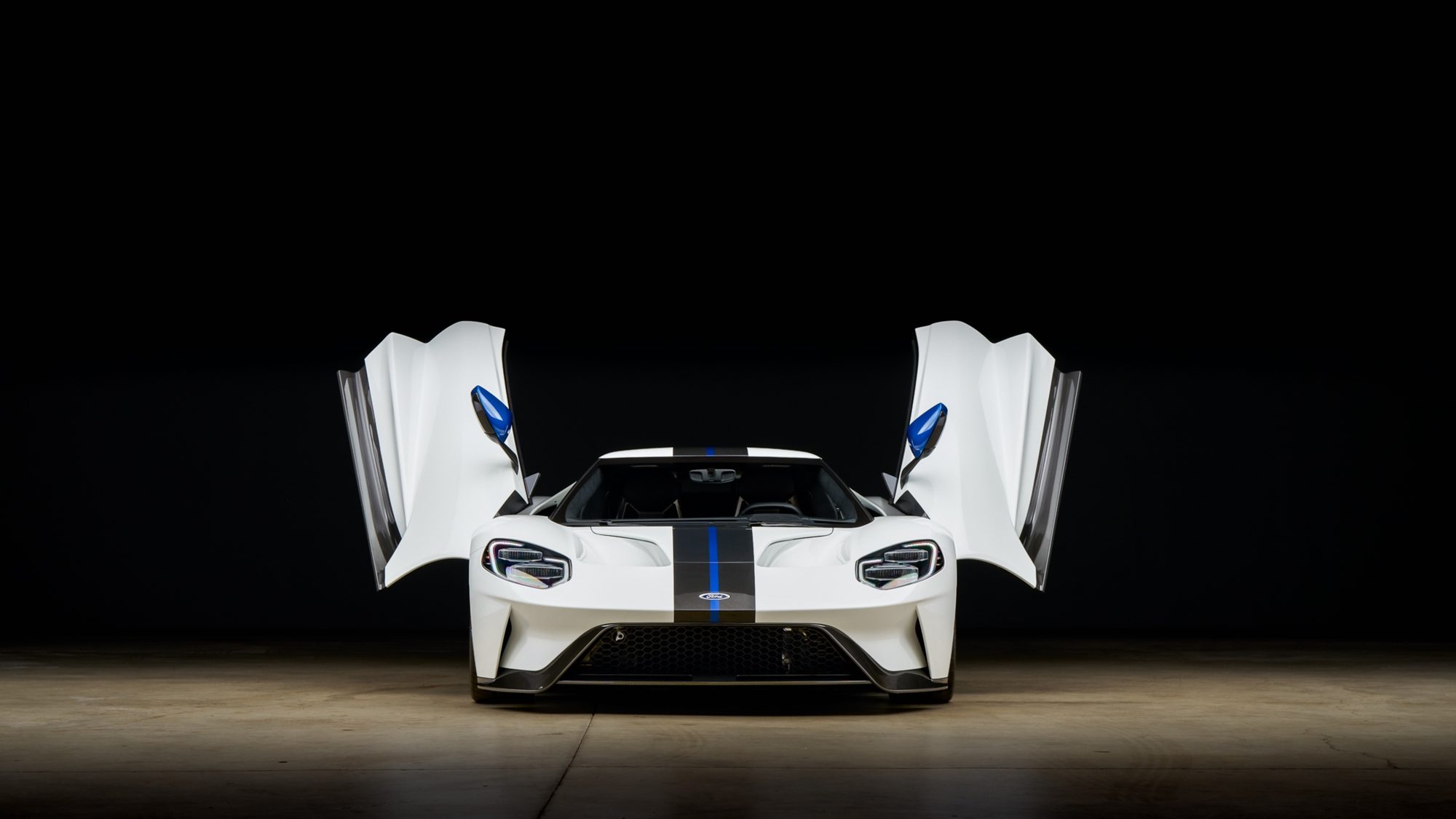 A white Ford GT seen head on in a dark studio, both doors raised, with blue mirror caps and a dark carbon fiber stripe with a thin blue center line running over the nose and roof