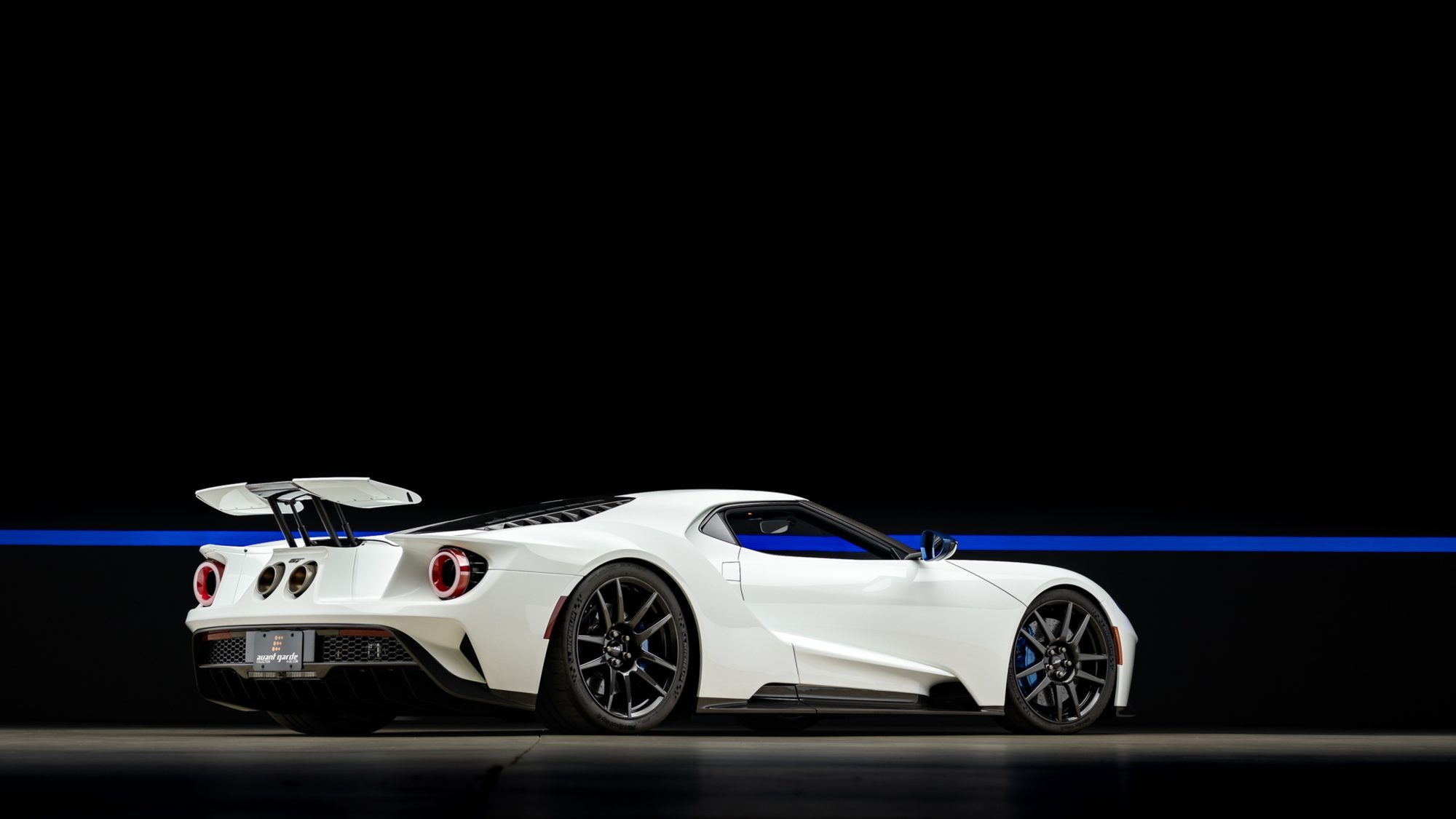 A white Ford GT from the rear three-quarter angle in a dark studio, its rear wing raised on struts, round red-ringed taillights and two center exhaust tips visible, a blue light line on the wall behind