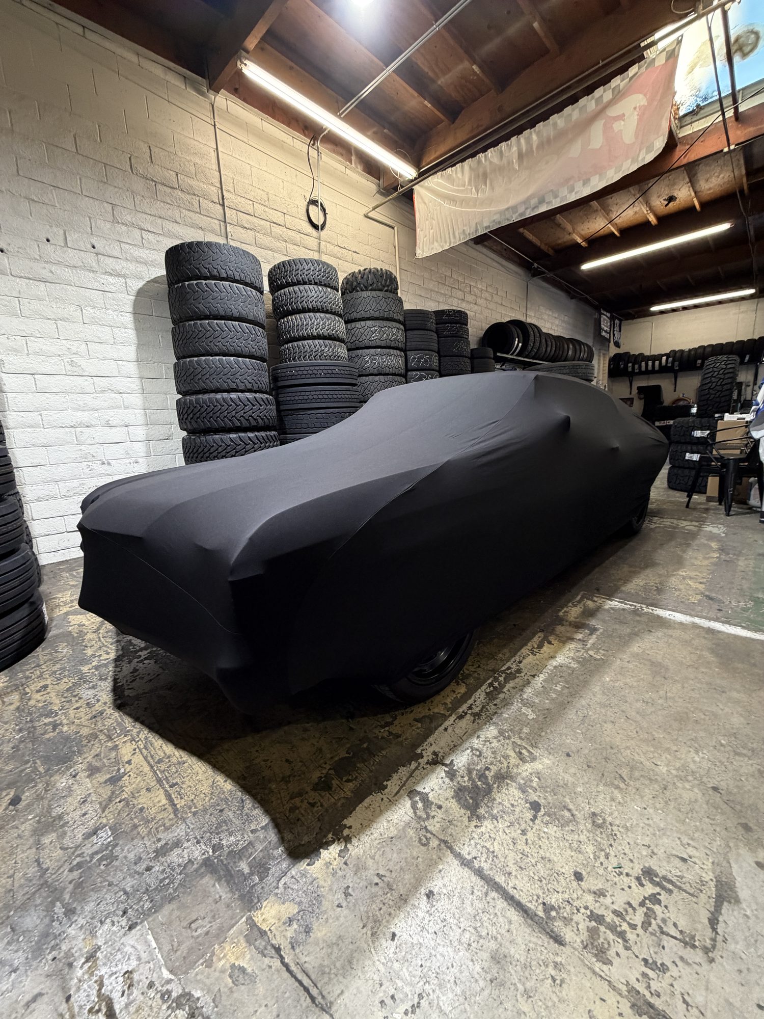 A classic muscle car under a fitted black stretch indoor cover in a tire shop, surrounded by stacks of tires