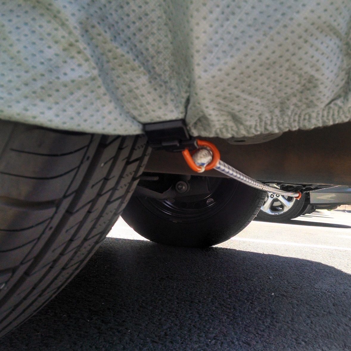 A close-up of a Gust Strap clip attached to the lower edge of a car cover, its cord running underneath the car toward the far side