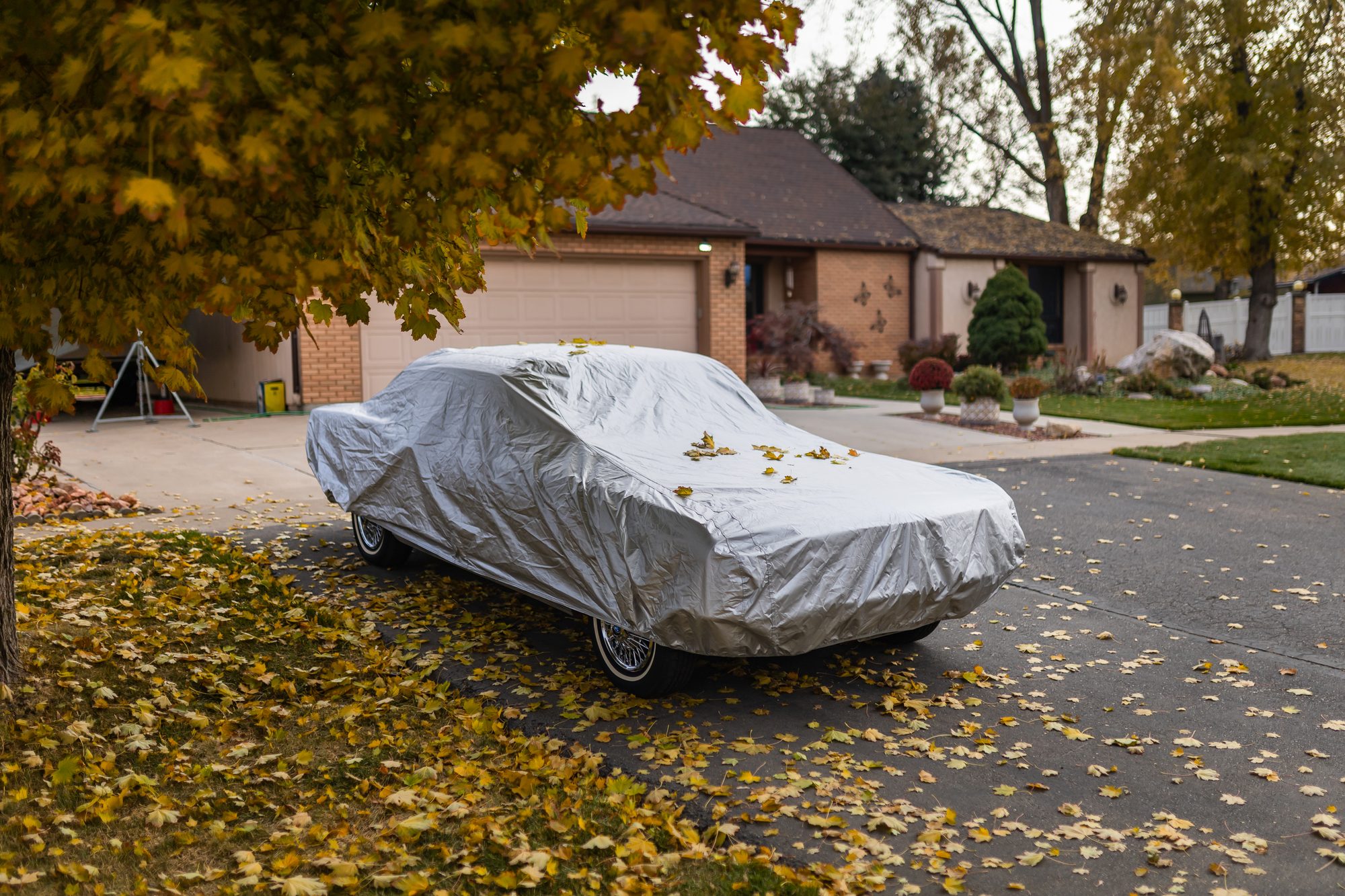 A classic car under a fitted silver outdoor cover on a driveway, with fallen autumn leaves on the cover and the pavement