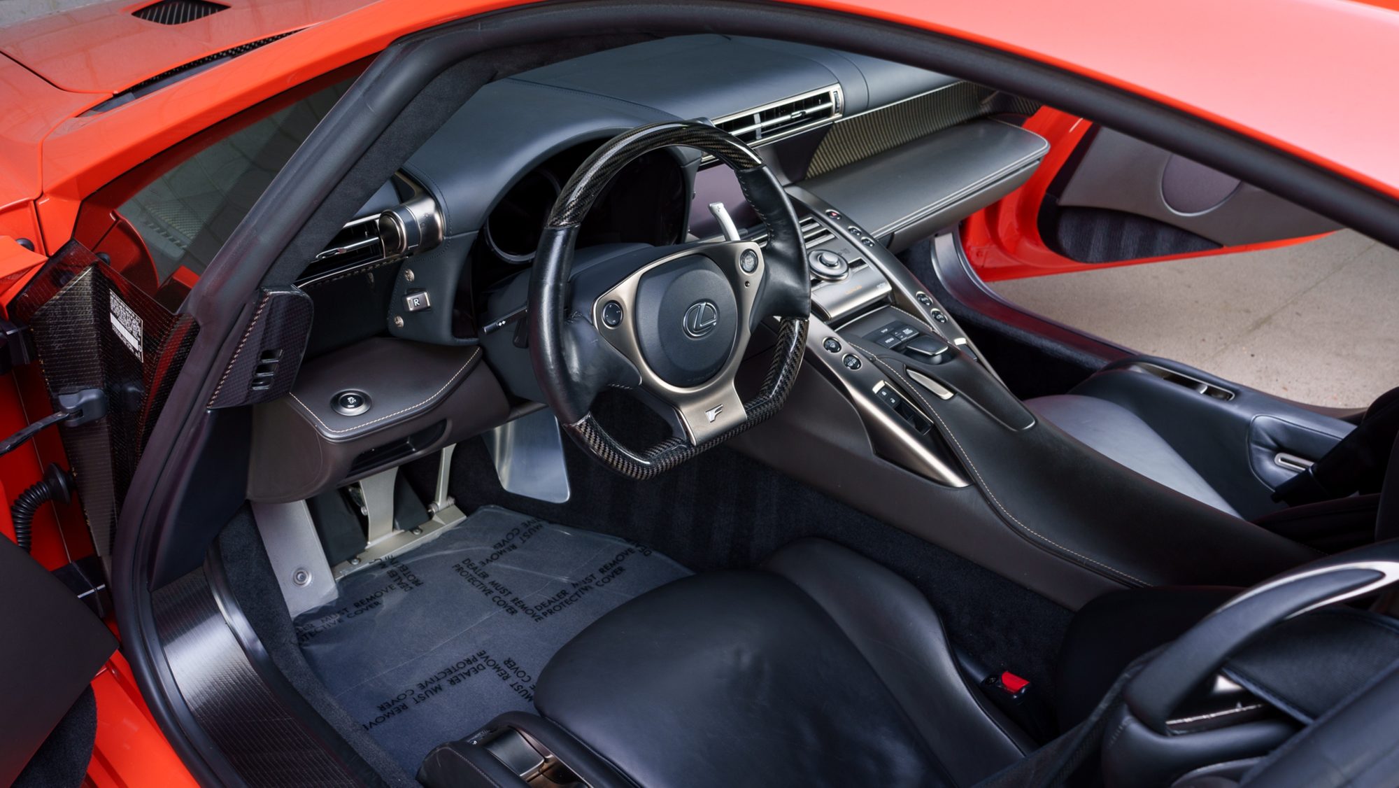 The Lexus LFA's black leather cockpit seen through the open driver's door, with a carbon-fiber and leather steering wheel, the center console, and the floor still under a dealer protective cover