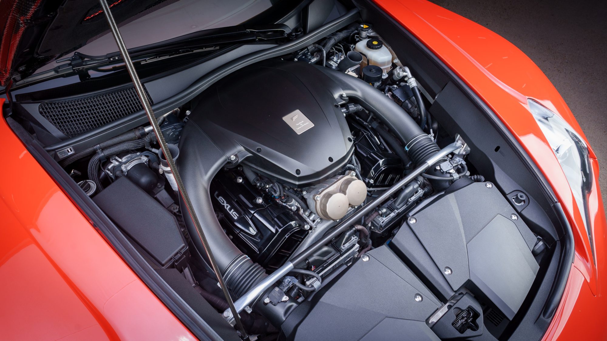 The Lexus LFA's V10 seen from above with the hood raised: a matte black engine cover with a small metal plaque reading Handbuilt by Lexus LFA Works, LEXUS lettering on the intake, and a prop rod holding the hood