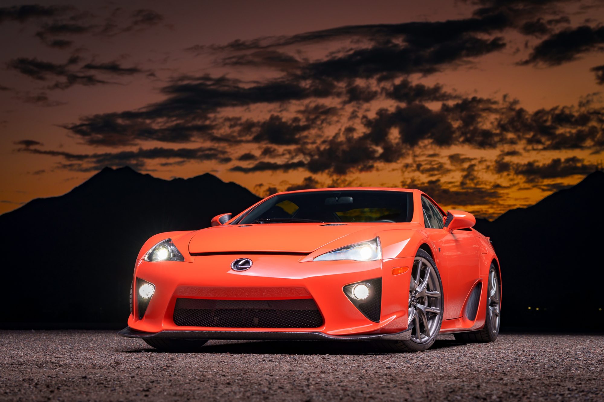 An orange Lexus LFA seen from the front three-quarter angle with its headlights on, parked on gravel under an orange sky with dark clouds and silhouetted mountains behind it