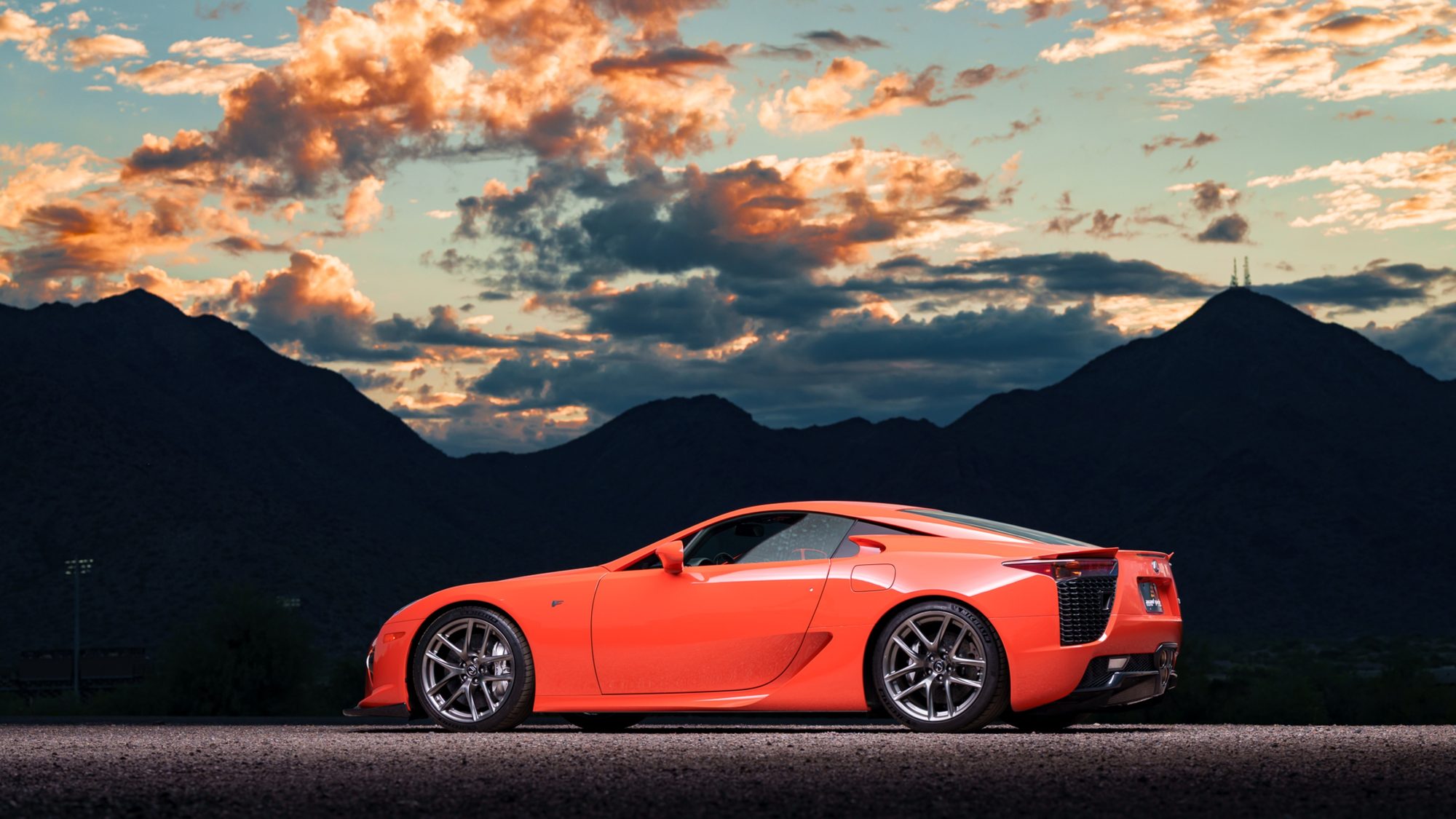 An orange Lexus LFA in side profile, parked on gravel in front of dark mountain silhouettes under a sky of orange-lit clouds