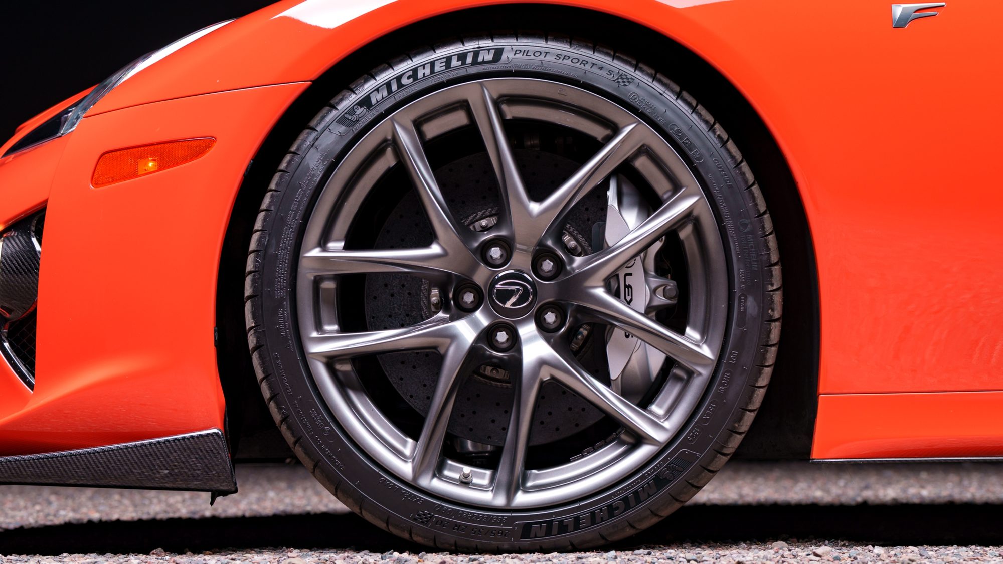 Close-up of an orange Lexus LFA's front wheel: a grey multi-spoke rim, a Michelin tire, a drilled brake disc and a silver caliper, with a chrome F badge on the fender above