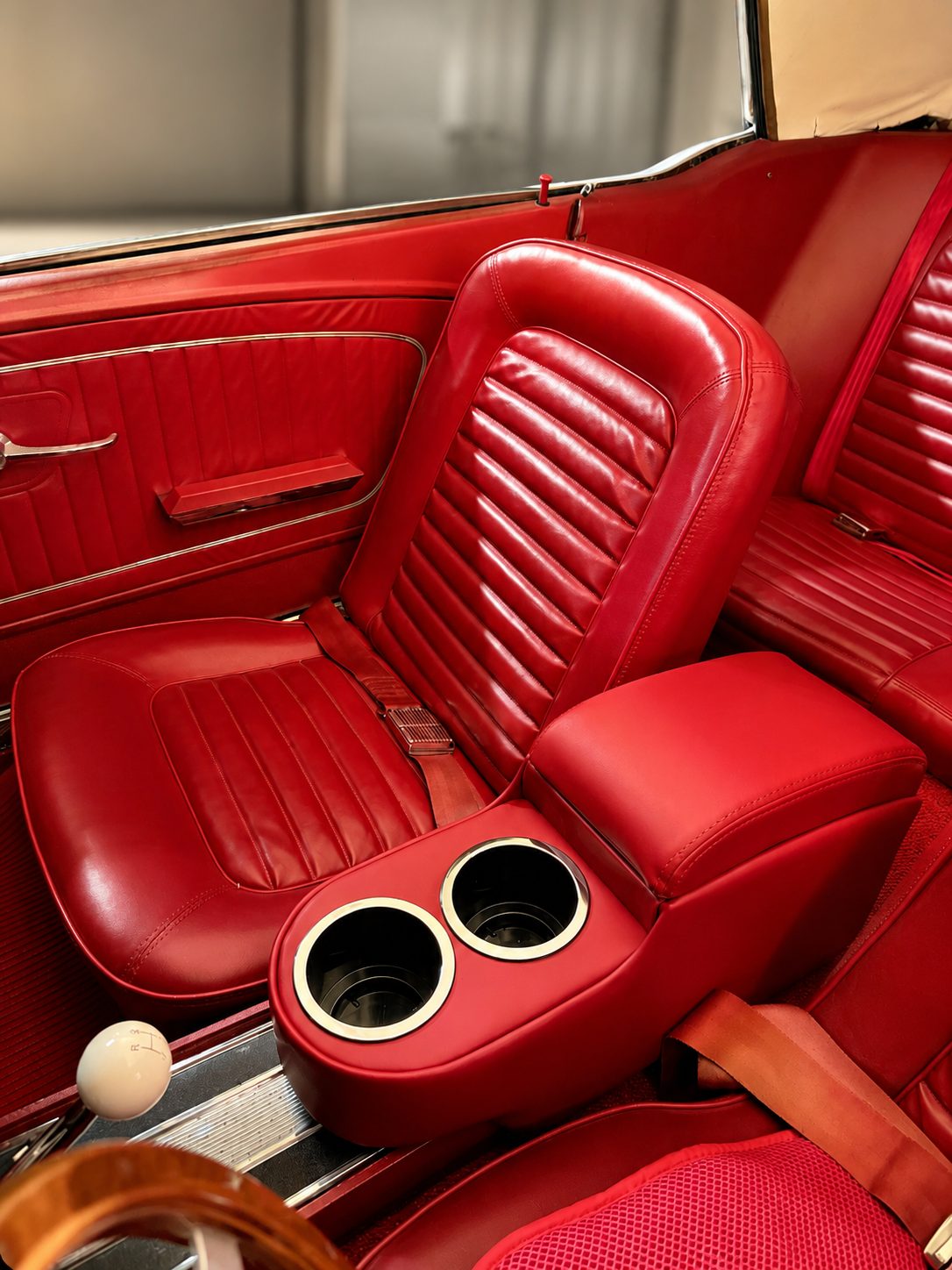 An illustration of a 1965 Mustang's red front bucket seat and center console with two cupholders, the red rear bench visible beyond