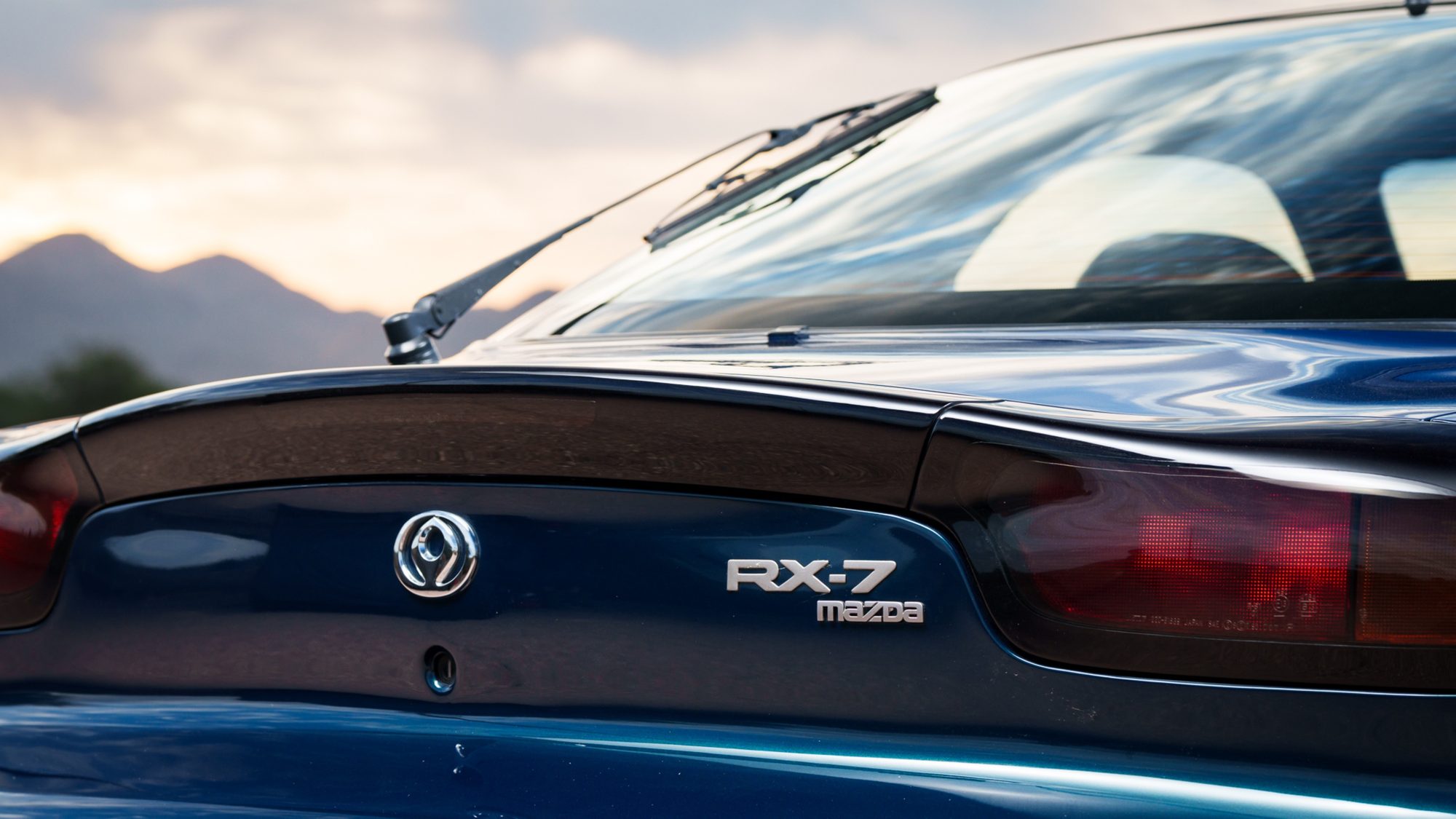 A close view of the RX-7 FD's tail, with the chrome Mazda emblem and the RX-7 and Mazda nameplate on dark blue paint below the rear spoiler and rear wiper