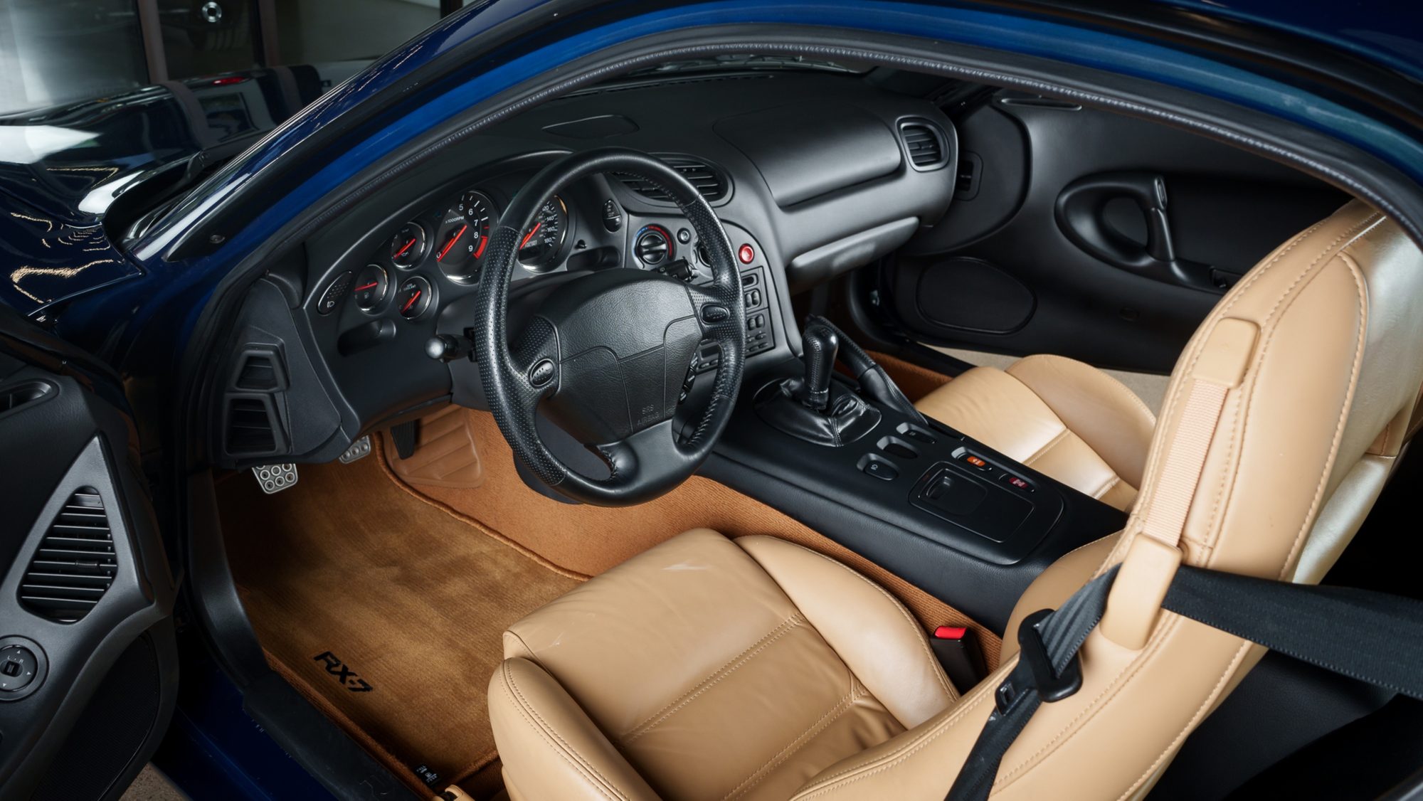 The Mazda RX-7 FD's black dashboard and tan leather seats seen through the open driver's door, with the leather steering wheel, round analog gauges and an RX-7 logo on the tan floor mat