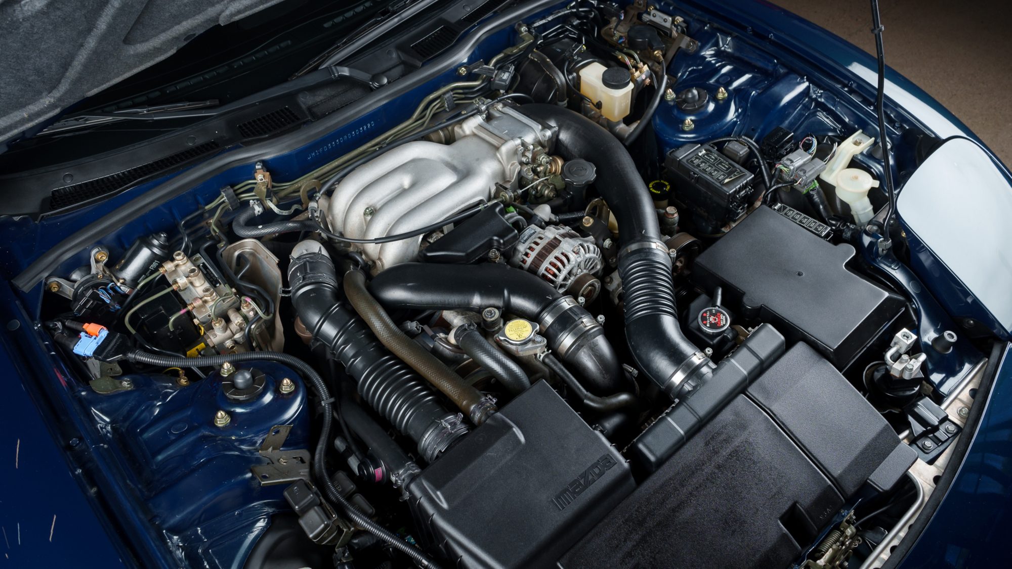 The Mazda RX-7 FD's engine bay seen from above, with the 13B-REW's silver intake manifold, black intake ducting and a black Mazda-embossed cover