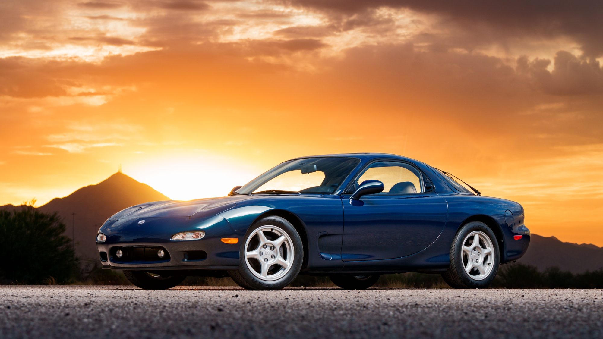A dark blue Mazda RX-7 FD seen from the front three-quarter angle on a desert road at sunset, with a mountain silhouette and glowing orange sky behind it