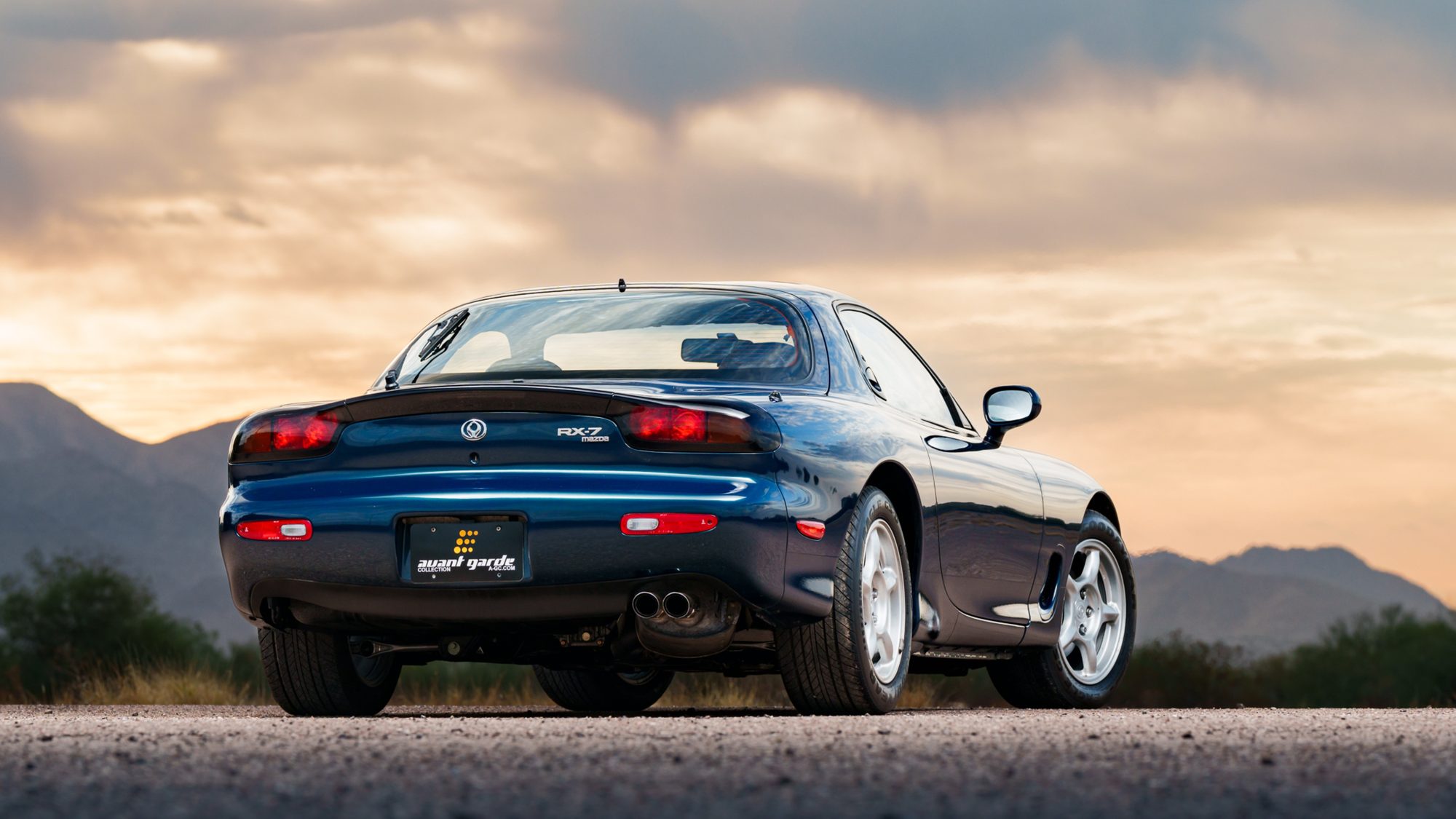 A dark blue Mazda RX-7 FD parked on pavement, seen from the rear three-quarter angle with mountains and an overcast sunset sky behind it