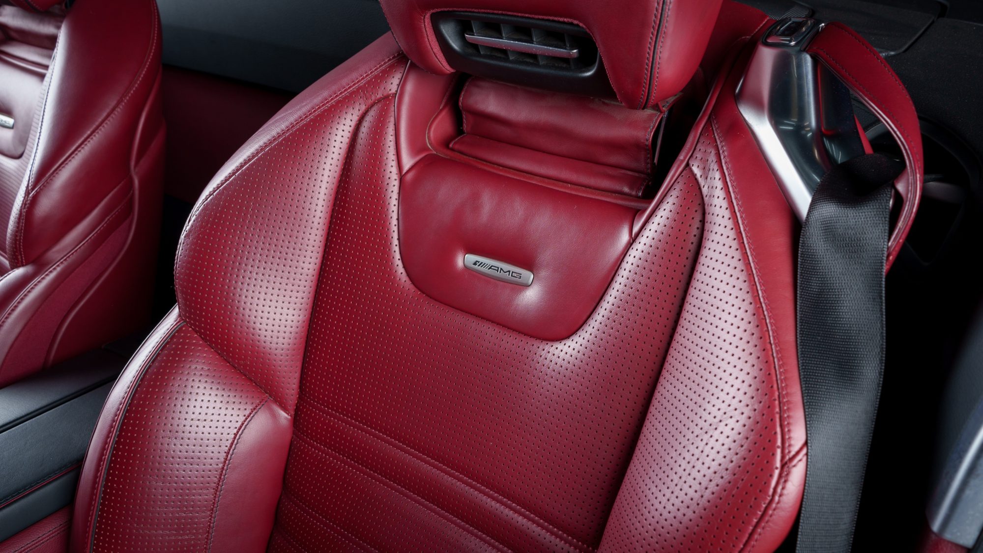 Close view of a red perforated-leather seatback with a metal AMG badge, an air vent in the headrest above it and a black seatbelt at right
