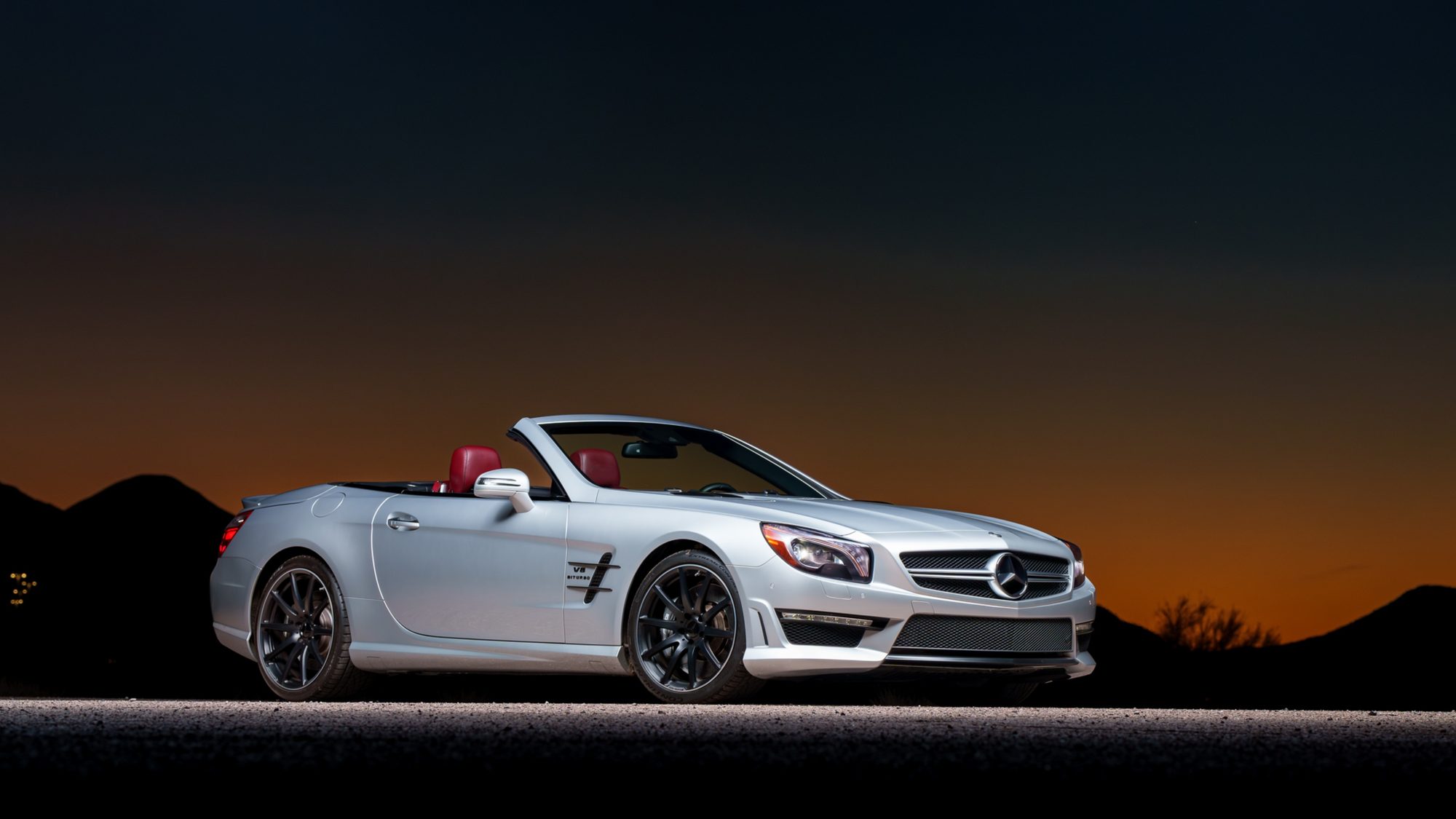 A silver Mercedes-Benz SL 63 AMG convertible with the top down and red leather interior, photographed from the front three-quarter angle at dusk with dark mountains and an orange gradient sky behind it