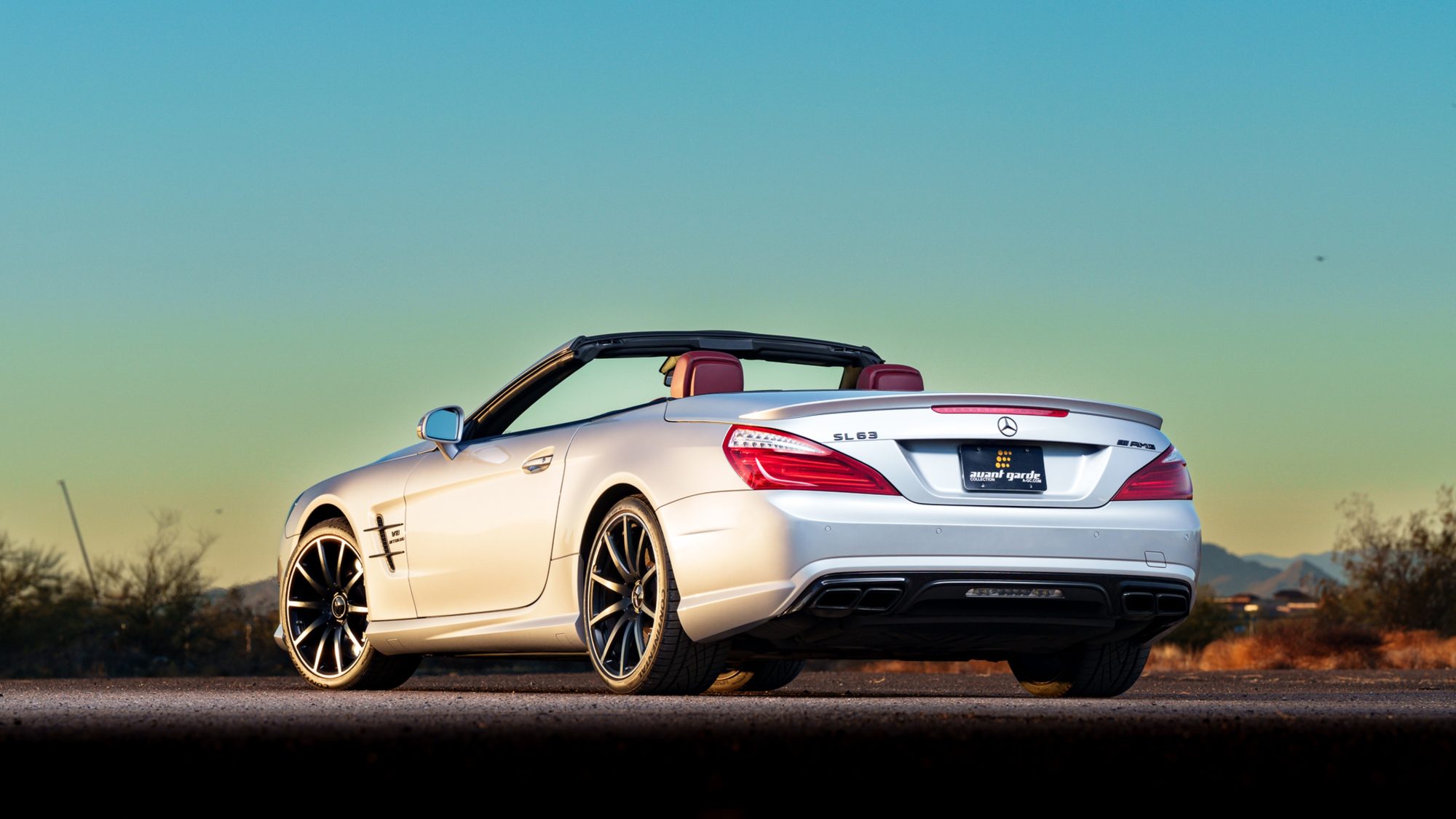 A silver Mercedes-Benz SL 63 AMG seen from the rear three-quarter angle with the top down, red seats and a dealer plate on the trunk lid, under a teal-to-gold sky with desert brush and hills behind it