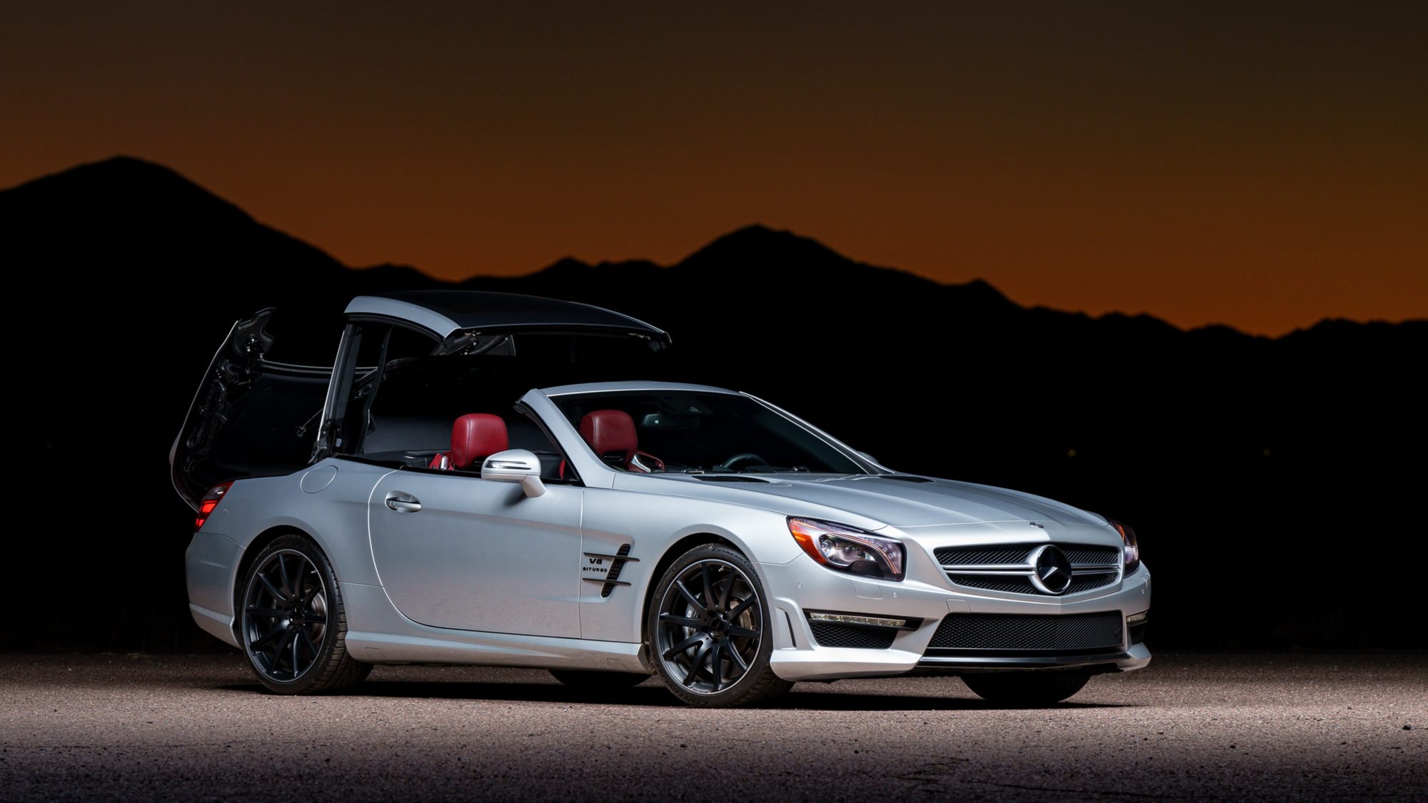 A silver Mercedes-Benz SL 63 AMG at dusk with its trunk lid swung up and back and the folding hardtop lifted above the cabin, partway through opening or closing, dark mountains behind it