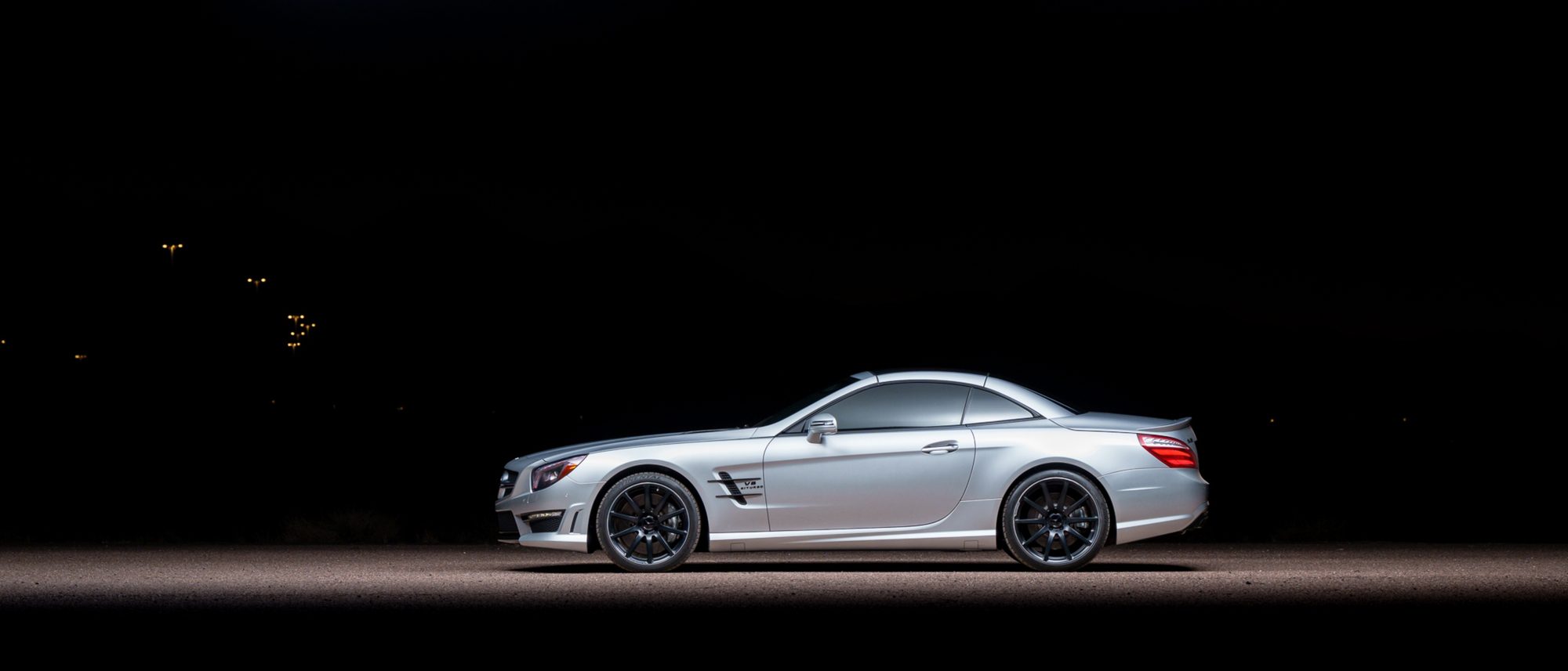 A silver Mercedes-Benz SL 63 AMG in side profile with its folding hardtop closed, dark wheels and a V8 Biturbo fender badge, lit against a black night background with a few distant lights at left