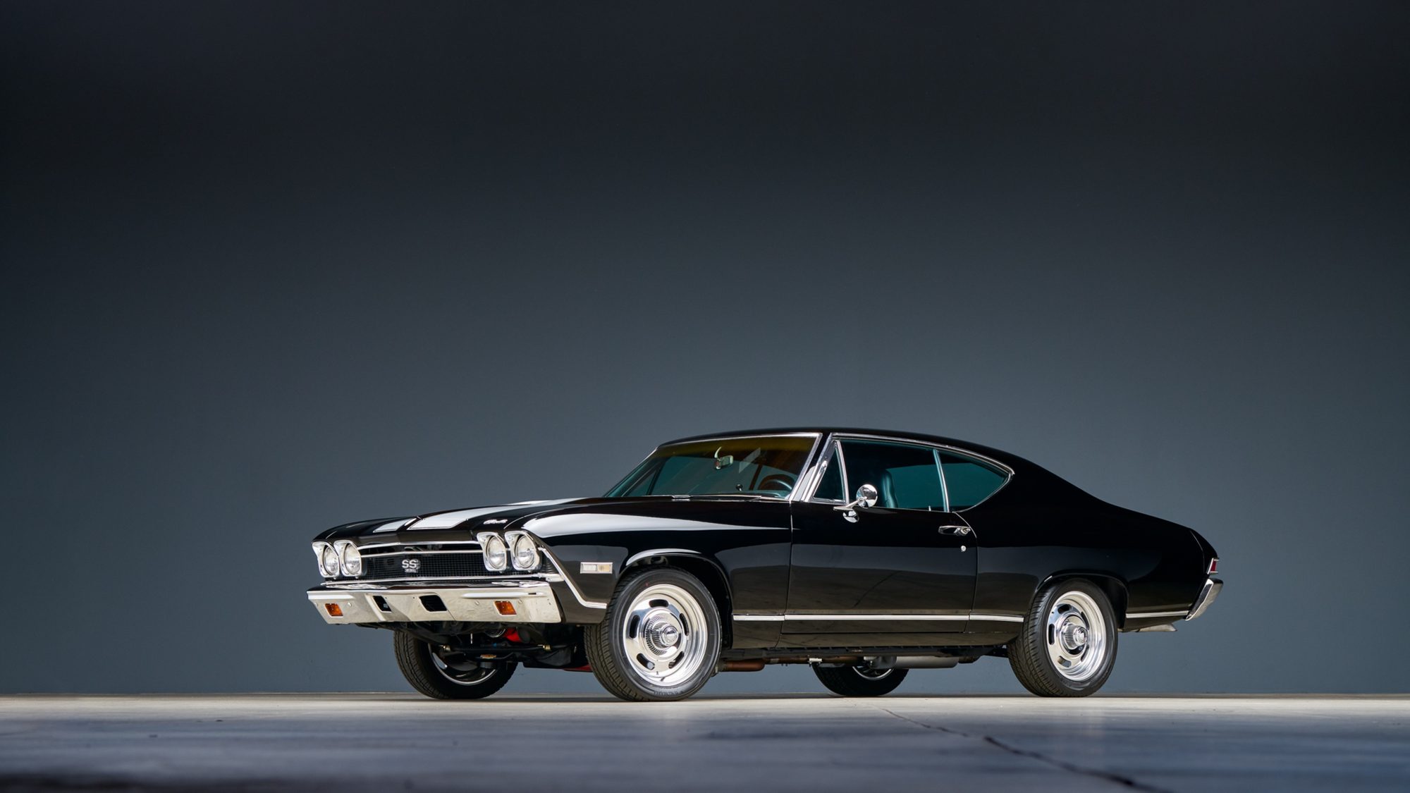A black Chevrolet Chevelle SS coupe with white stripes on the hood, photographed from the front three-quarter angle in a dark studio