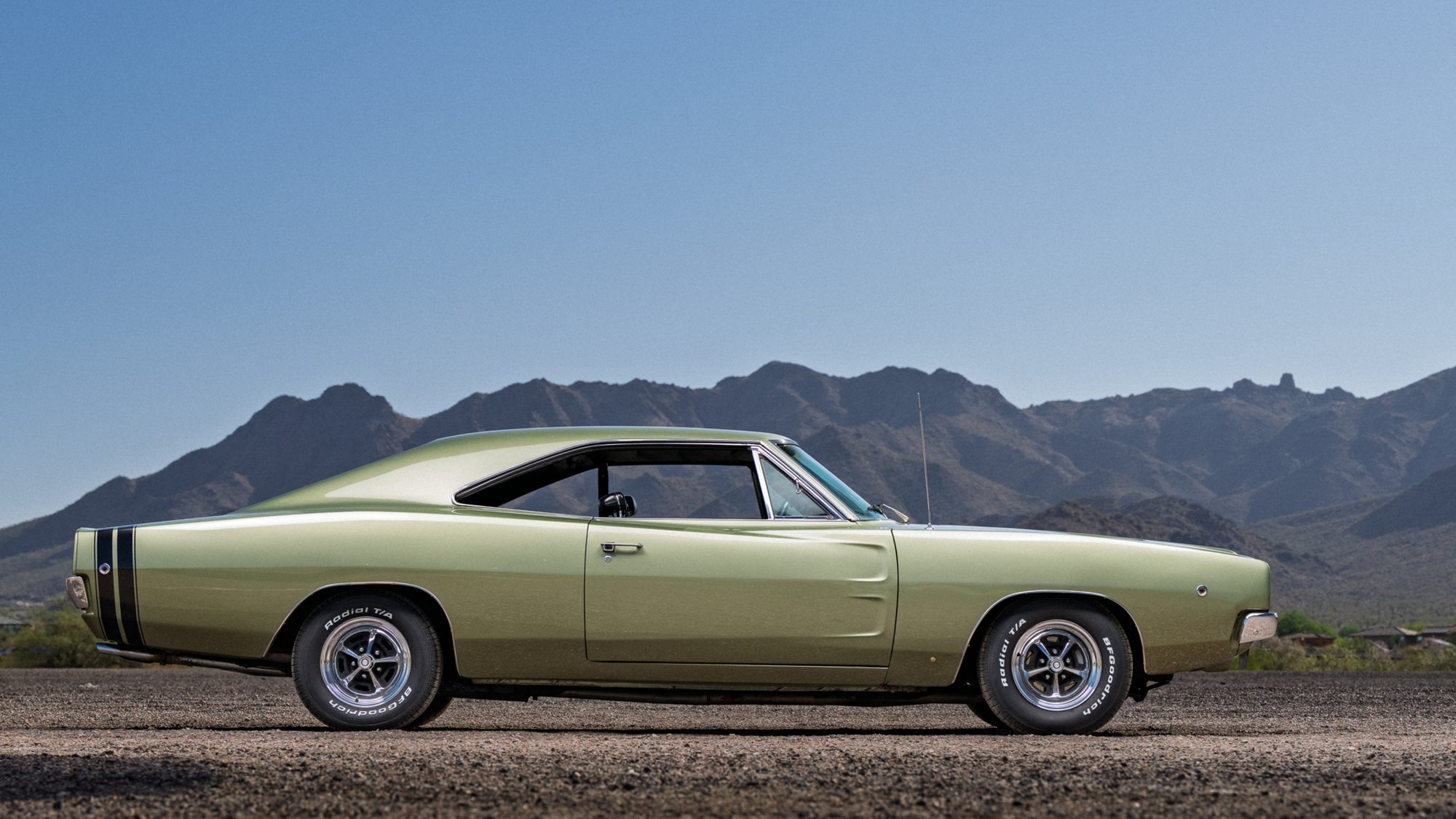 A light green metallic Dodge Charger hardtop in profile, with twin black stripes wrapped around its tail, parked on gravel in front of desert mountains