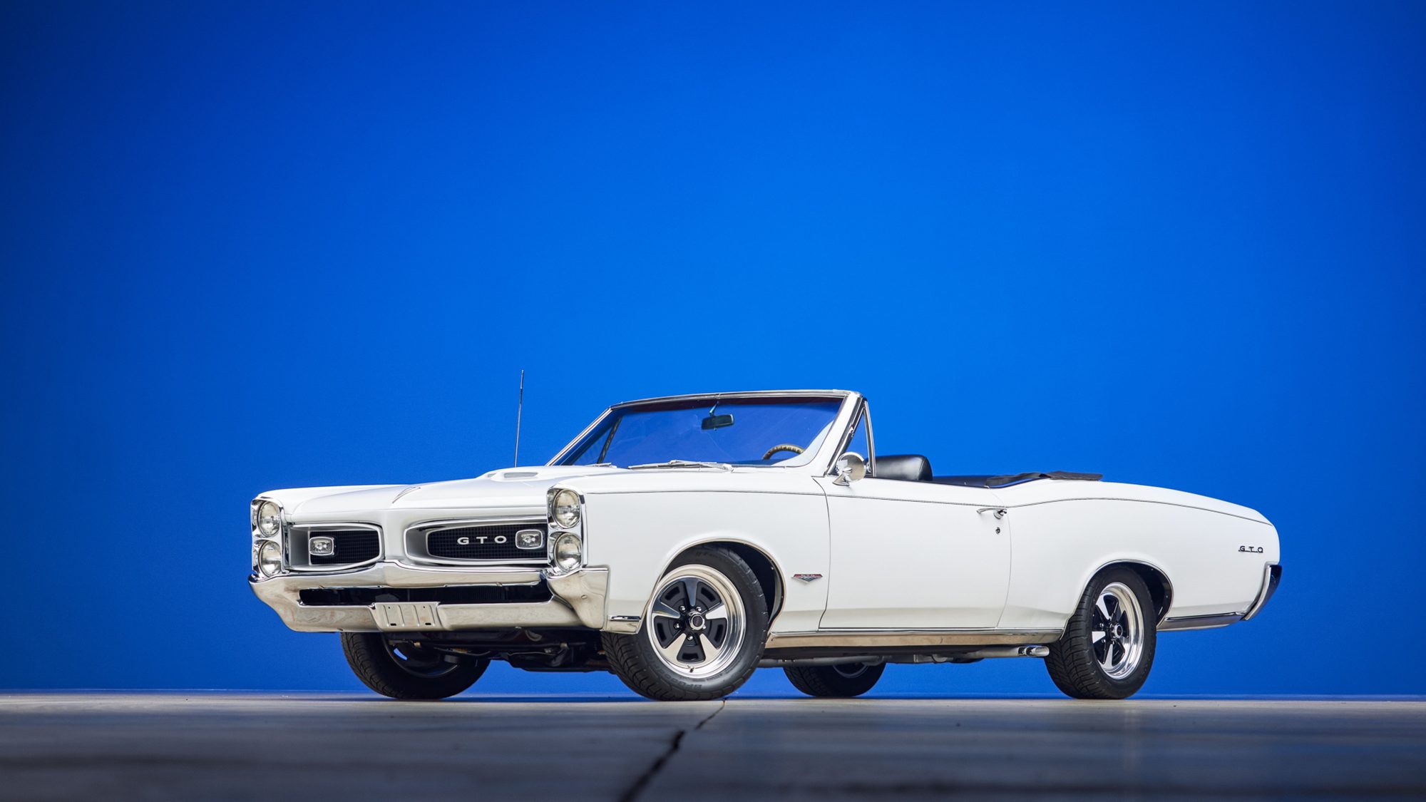 A white Pontiac GTO convertible with its top down, photographed from the front three-quarter angle against a solid blue background