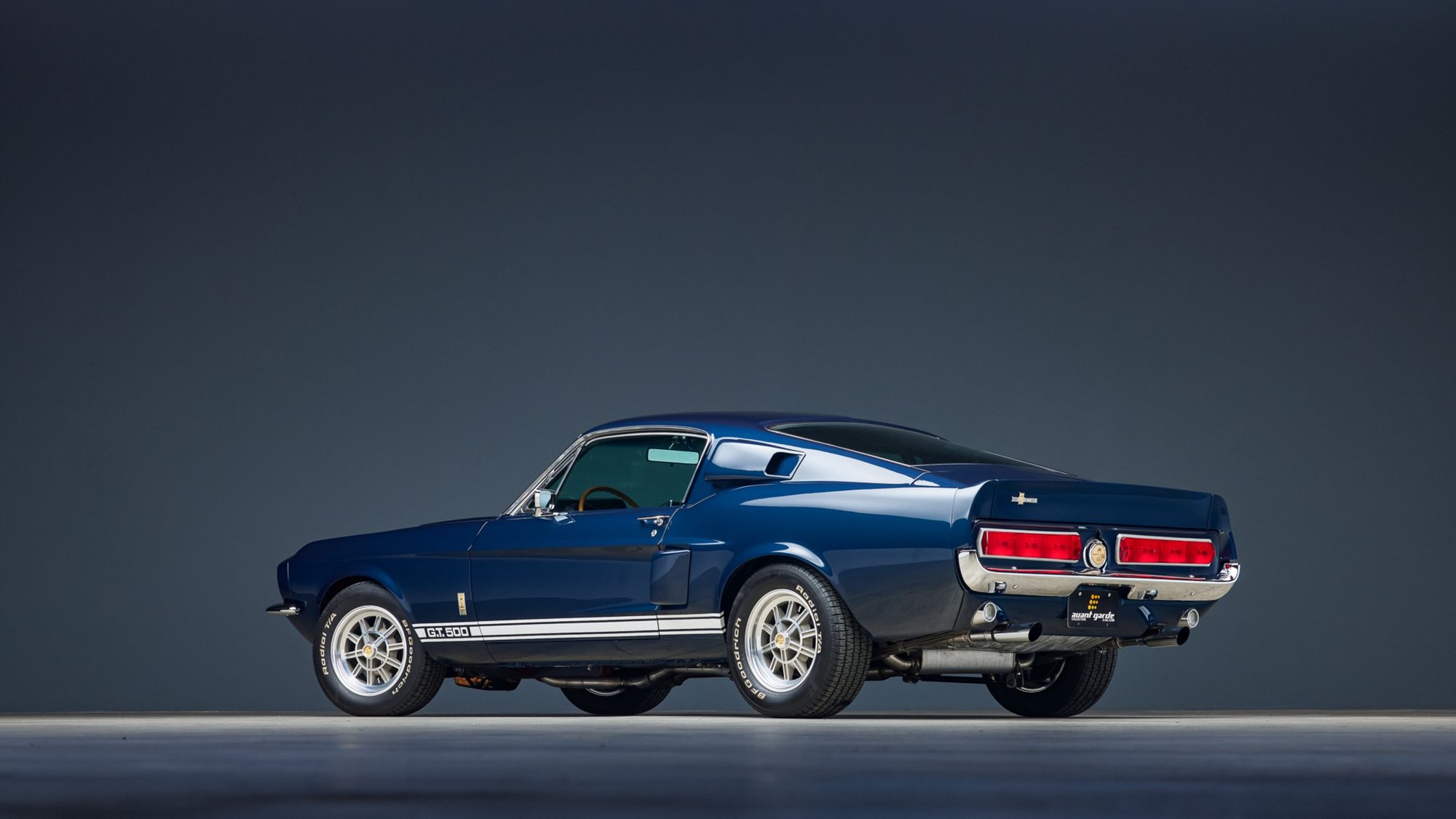 A dark blue Shelby GT500 fastback seen from the rear three-quarter angle in a dark studio, with white rocker stripes reading G.T. 500 and twin exhaust tips
