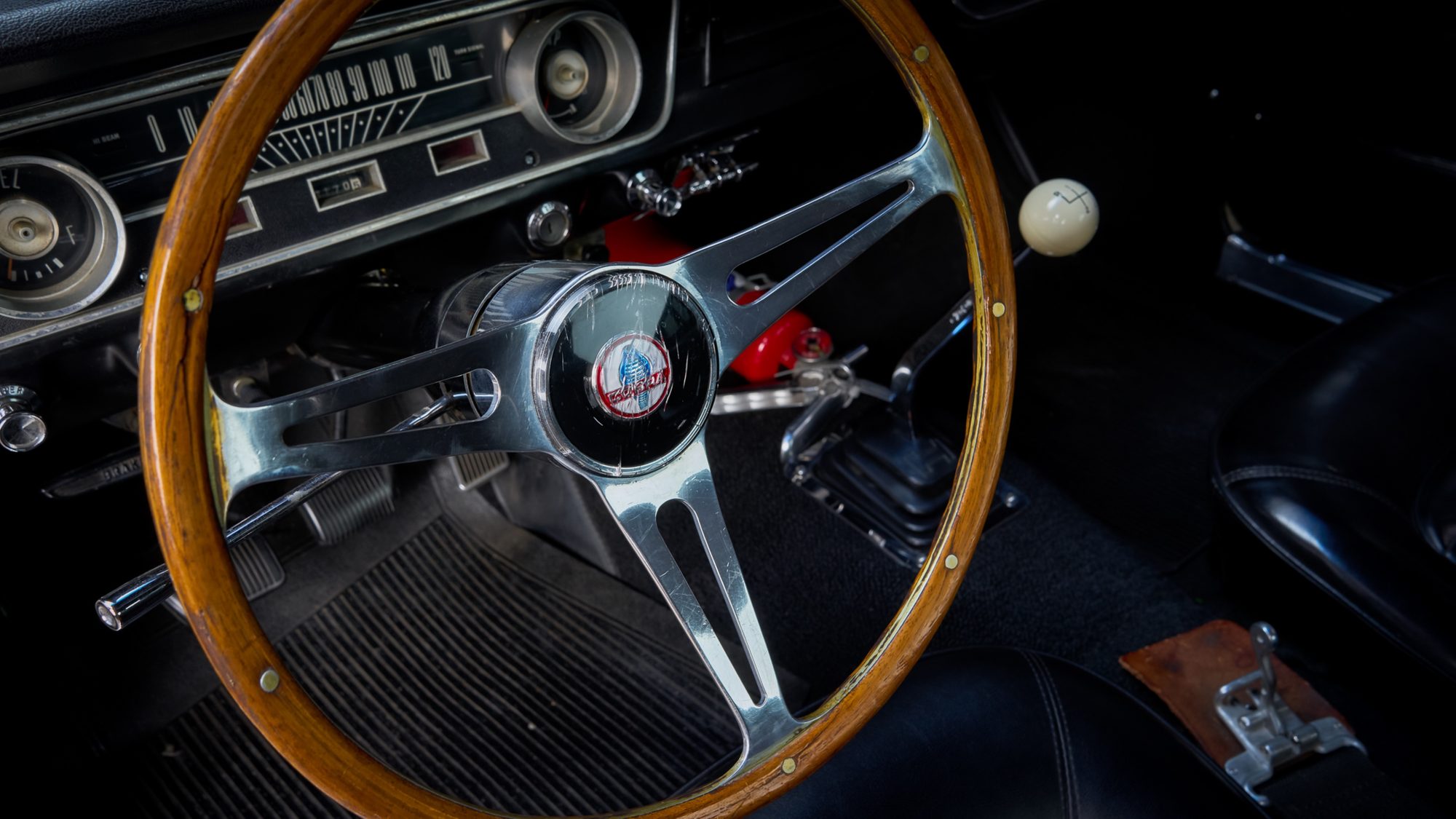 A wood-rimmed steering wheel with a Cobra horn button in front of an early Mustang's horizontal speedometer, with a white shift knob on the floor-mounted four-speed shifter