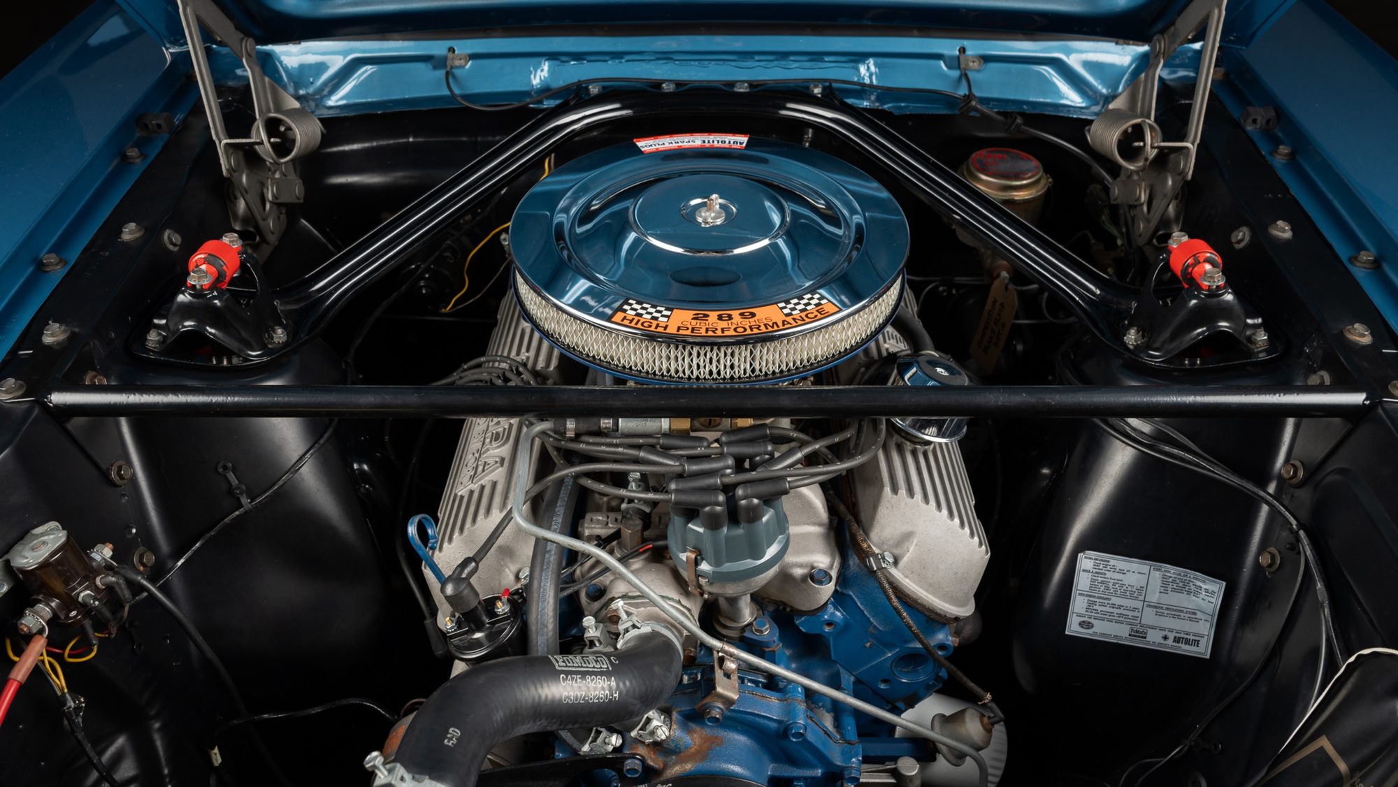 The engine bay of a blue Shelby GT350: a chrome air cleaner with a 289 High Performance decal, finned aluminum valve covers, and black braces linking the shock towers to each other and to the firewall
