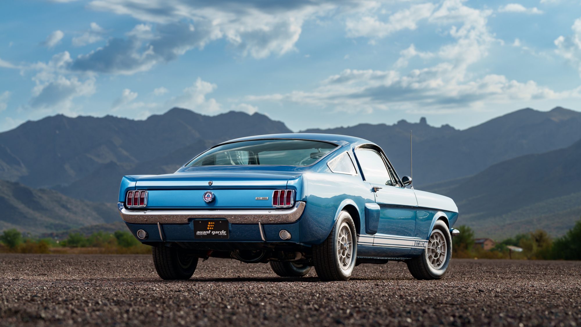 A blue Shelby GT350 fastback seen from the passenger-side rear three-quarter angle, parked on gravel under a cloudy sky with desert mountains behind it
