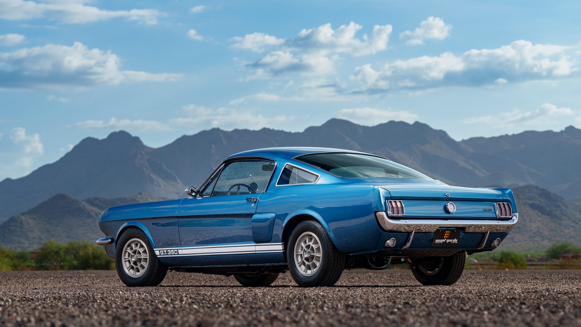 A blue Shelby GT350 fastback seen from the driver-side rear three-quarter angle, with G.T. 350 on its rocker stripe and desert mountains behind it