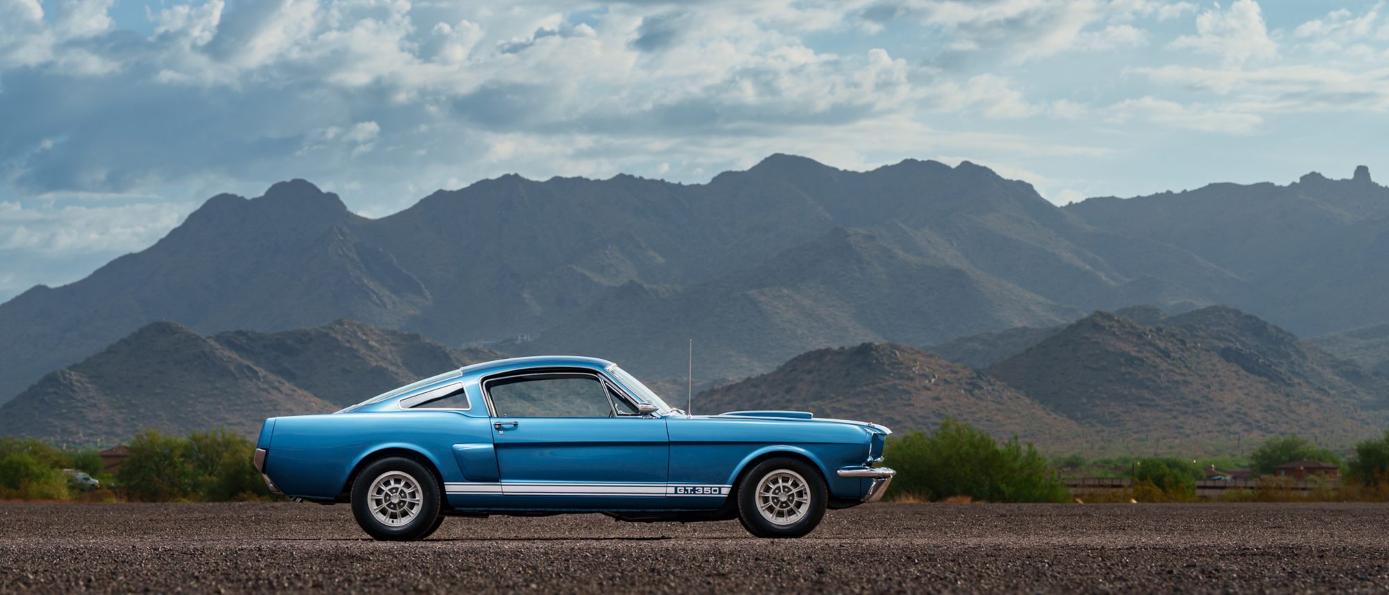 A blue Shelby GT350 fastback in side profile on gravel, desert mountains behind it, showing its rear-quarter window, side scoop and G.T. 350 rocker stripe