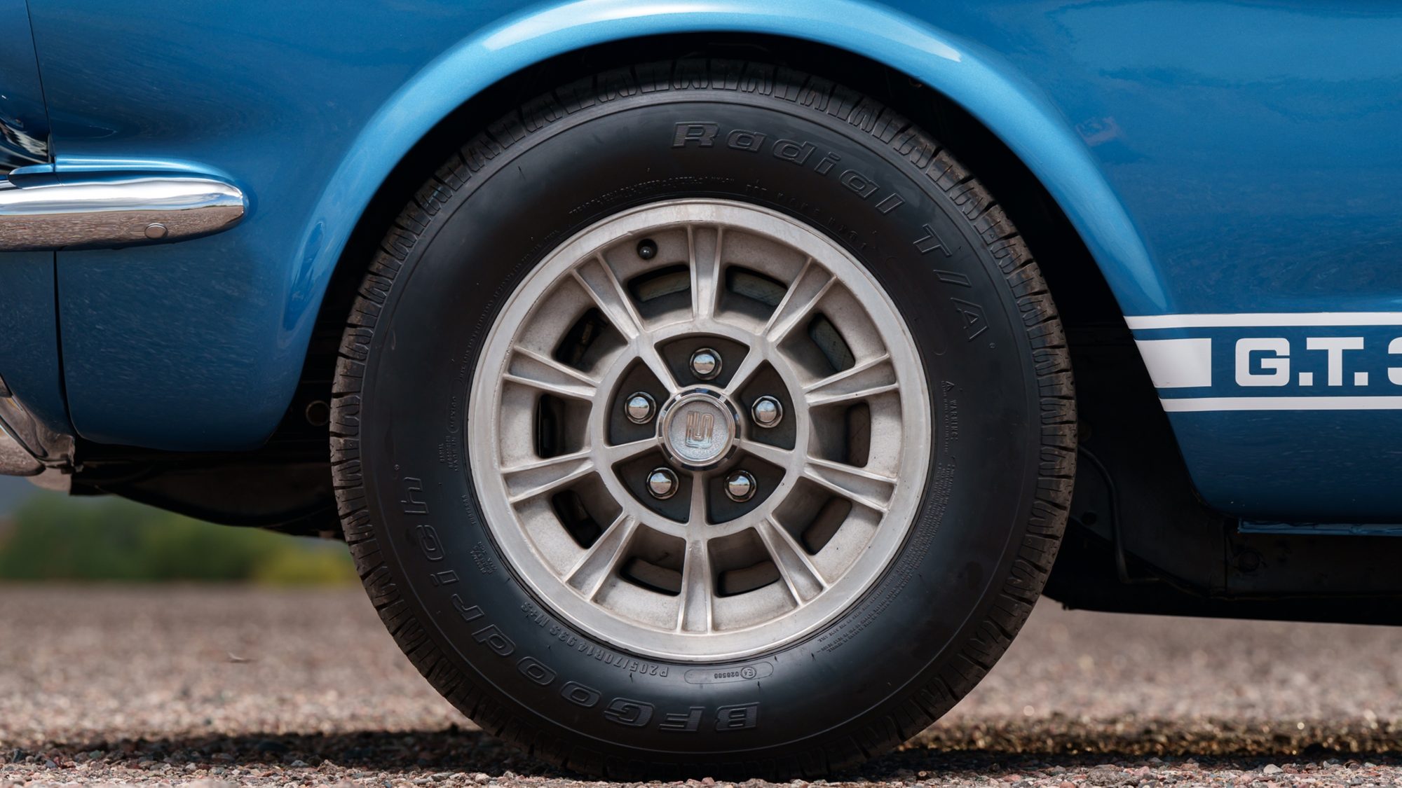 The front wheel of a blue Shelby GT350, a silver multi-spoke alloy, with G.T. 350 lettering on the white rocker stripe behind it