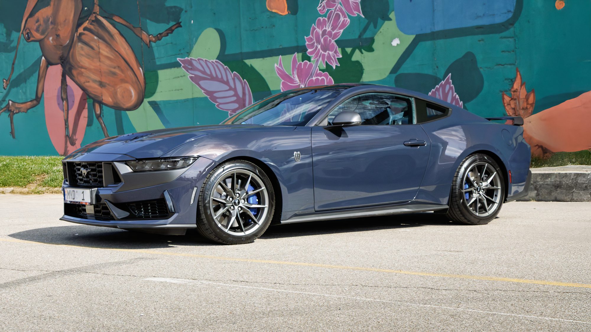 A dark blue 2024 Ford Mustang Dark Horse parked in front of a colorful painted mural of a beetle and pink flowers, seen from the front three-quarter angle