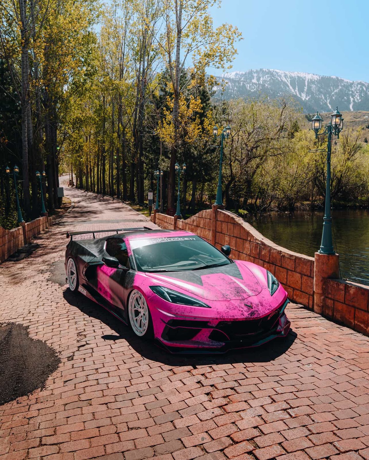 The pink and black wrapped Corvette, with a tall rear wing, parked on a brick path beside a pond, lined with trees and lamp posts, a snow-capped mountain in the distance