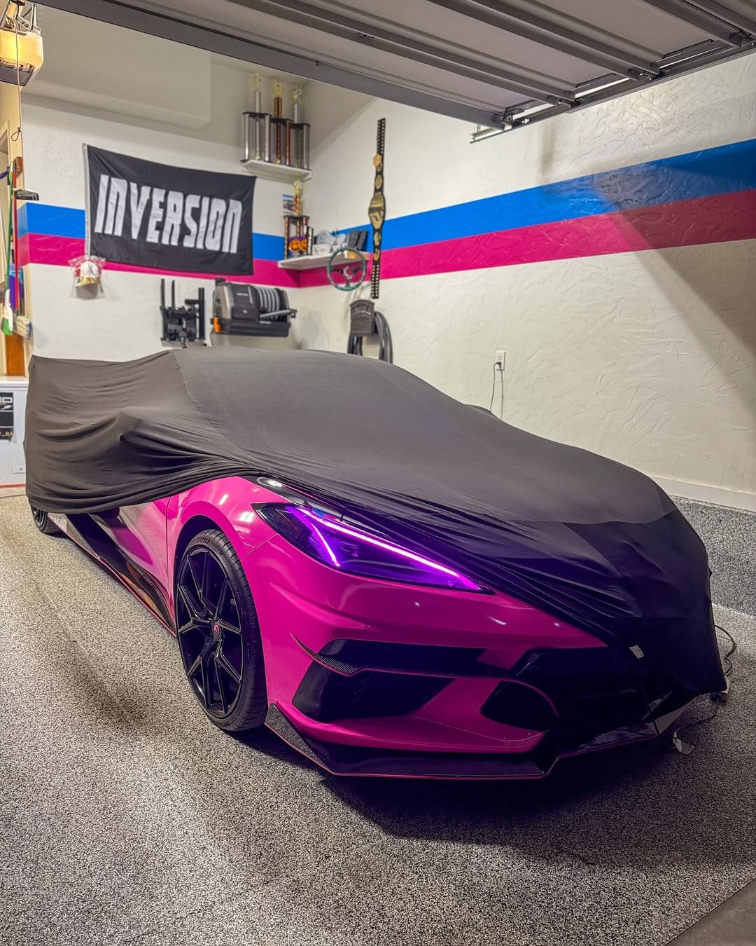 A black car cover pulled most of the way over the Corvette in a home garage, leaving its gloss pink front bumper, a black wheel and a purple-lit headlight showing