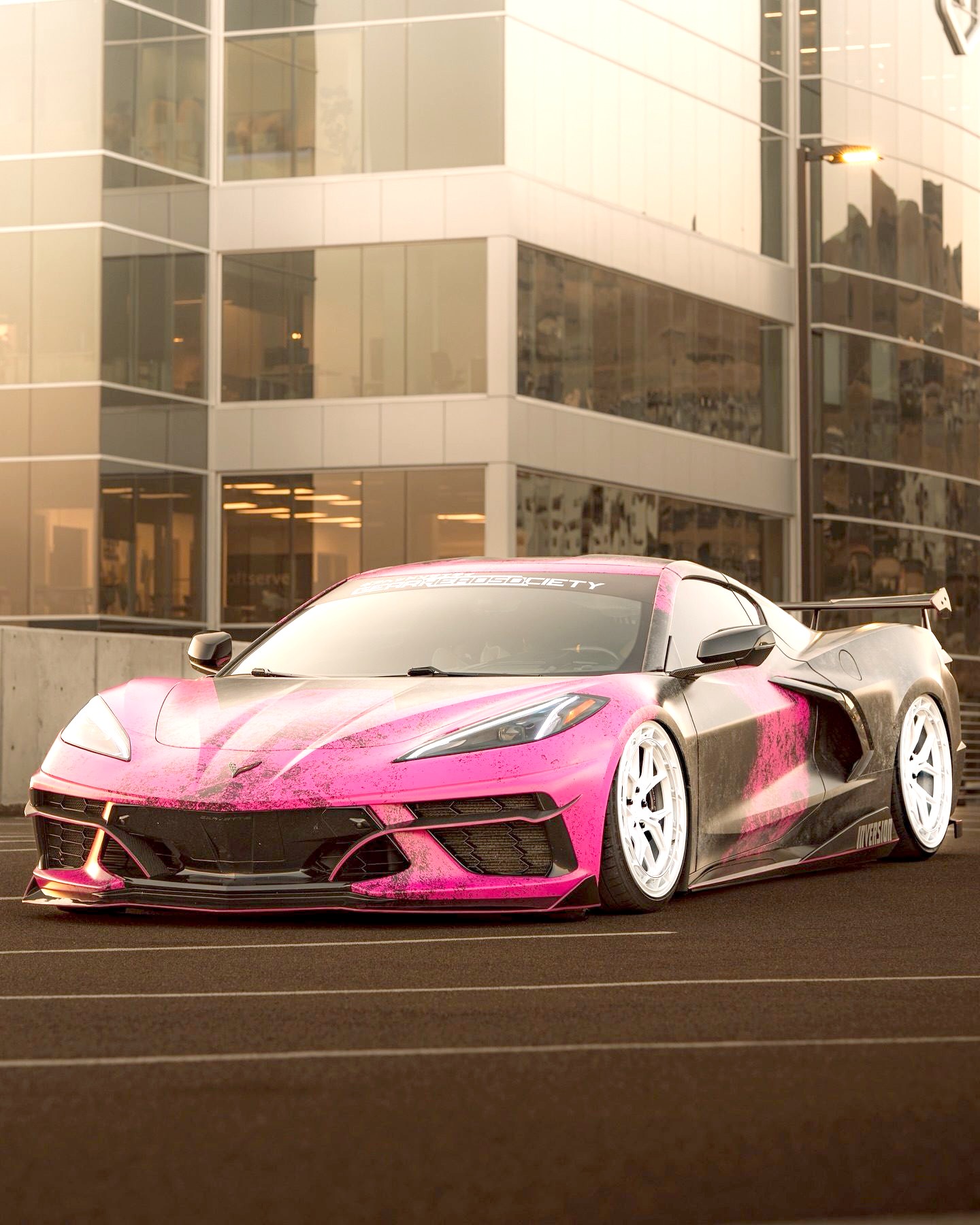 A pink and black distressed-wrap Corvette Stingray on white wheels, parked front three-quarter in front of a glass office building in evening light, a Gearhead Society banner across the top of its windshield