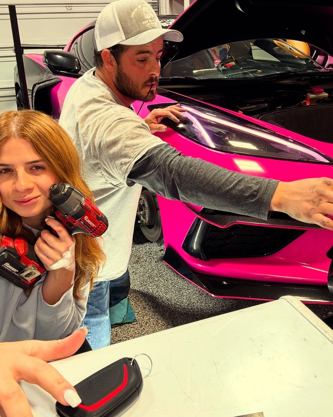 A young woman holds a cordless impact driver while a man in a cap works at the front of a gloss pink Corvette in a home garage, its front trunk open