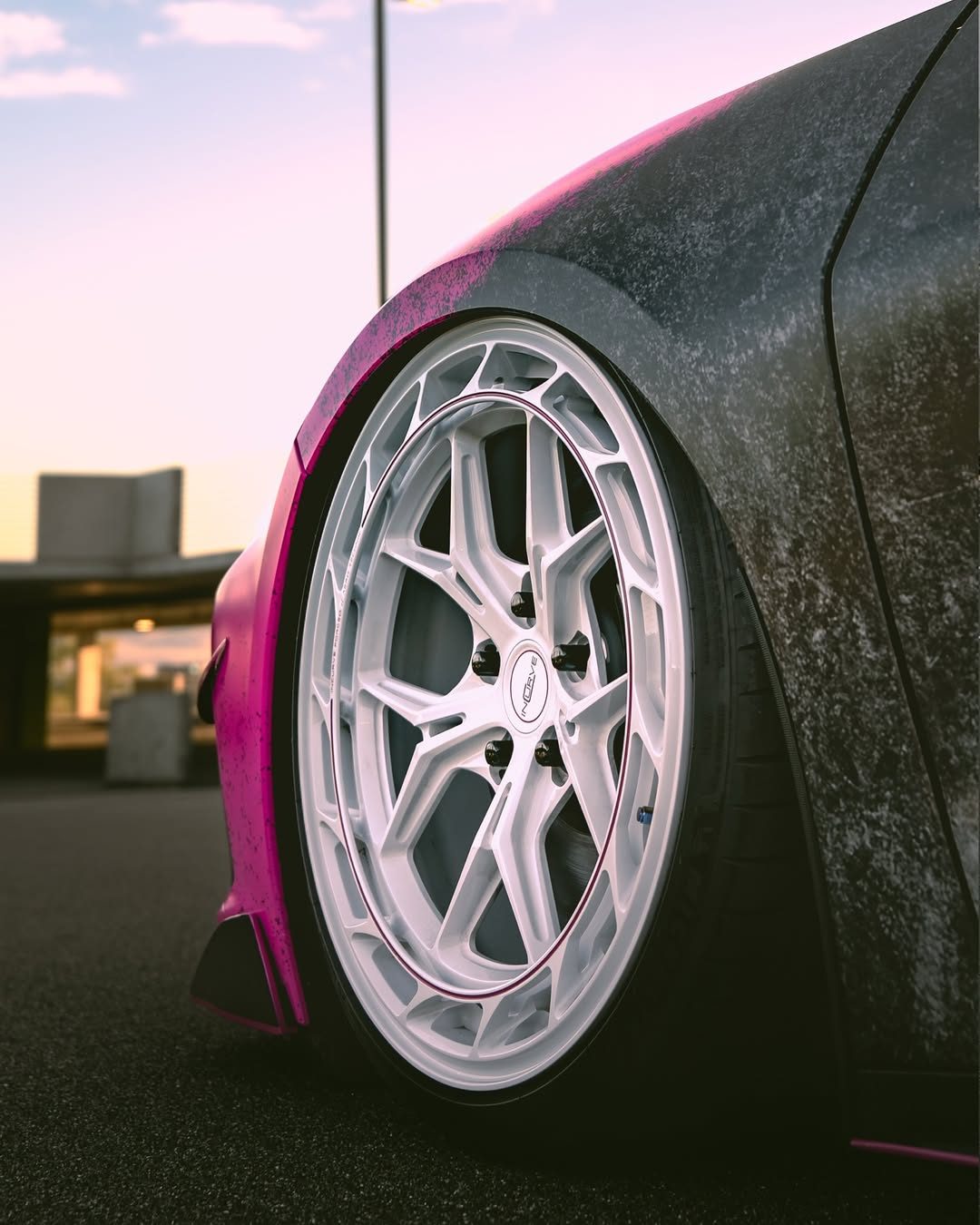A close-up of a white multi-spoke wheel tucked into the Corvette's fender, the pink and black distressed wrap on the bodywork around it, under an evening sky
