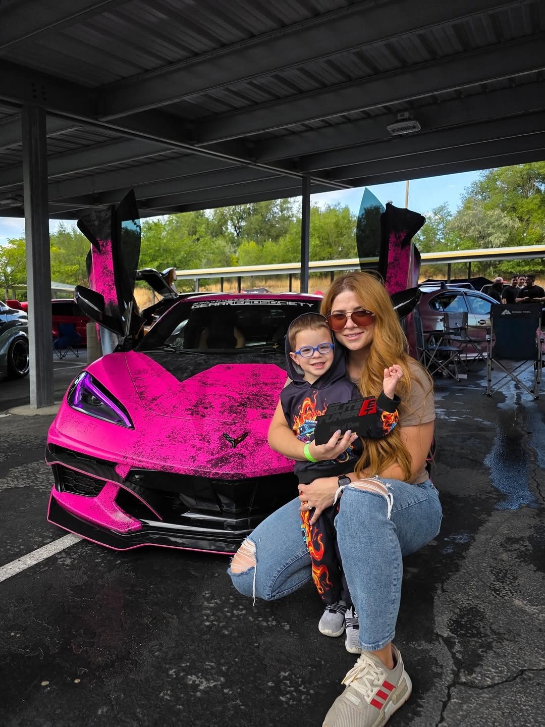A woman in sunglasses kneels under a carport at a car show with a young boy in a flame-print hoodie, in front of the pink and black wrapped Corvette Stingray with its doors raised; she holds a black Elite Tuner award plaque in front of the boy