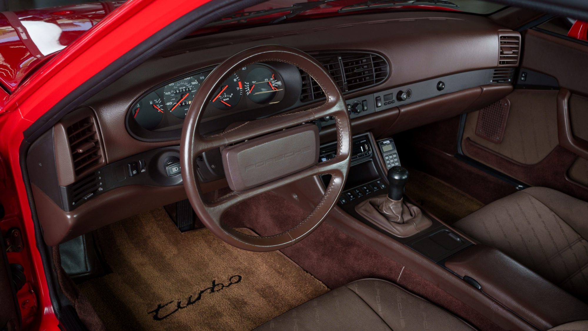 The brown interior of a Porsche 944 Turbo: analog gauges, a four-spoke steering wheel, cloth seats and a floor mat embroidered turbo