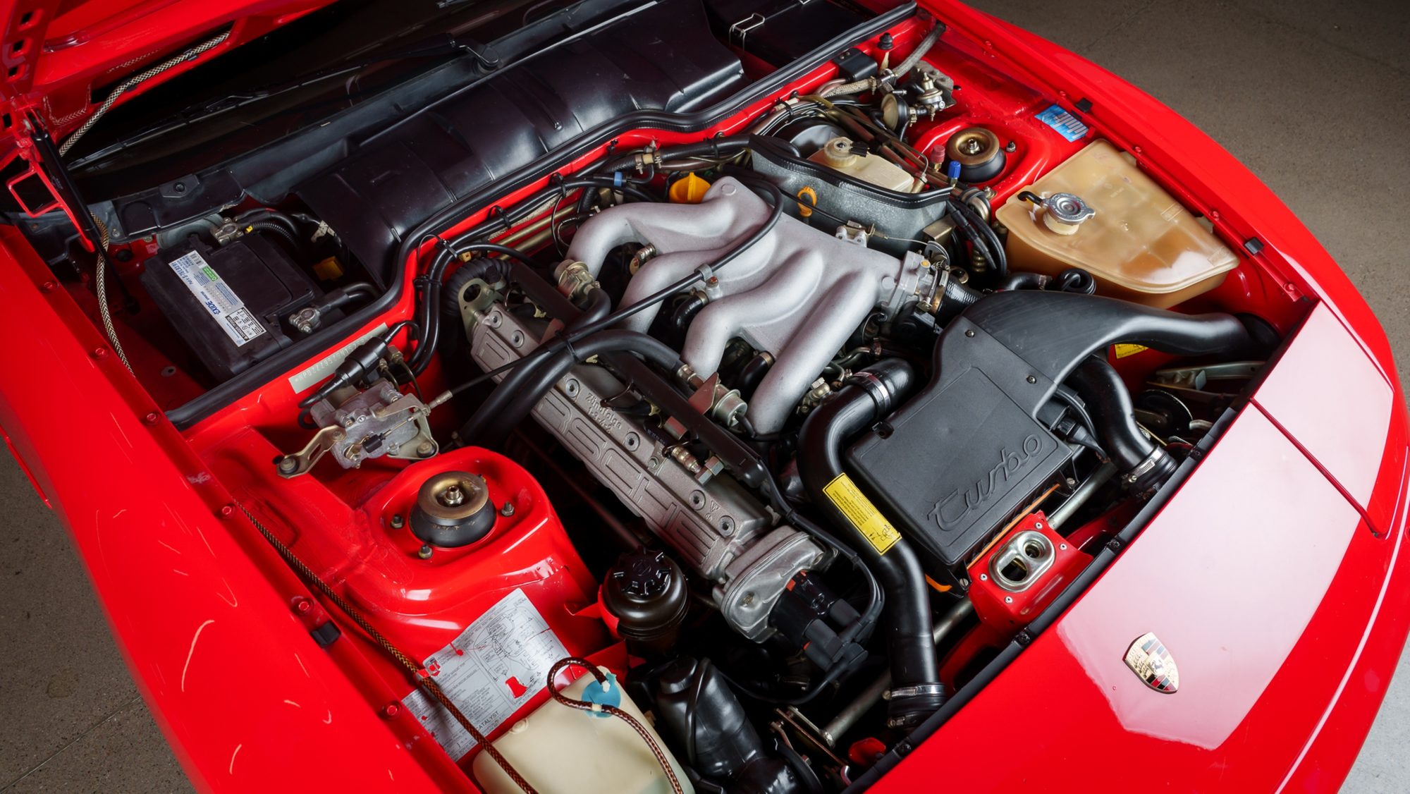 The engine bay of a red Porsche 944 Turbo, with the four-cylinder's cam cover, silver intake manifold and a black housing marked turbo