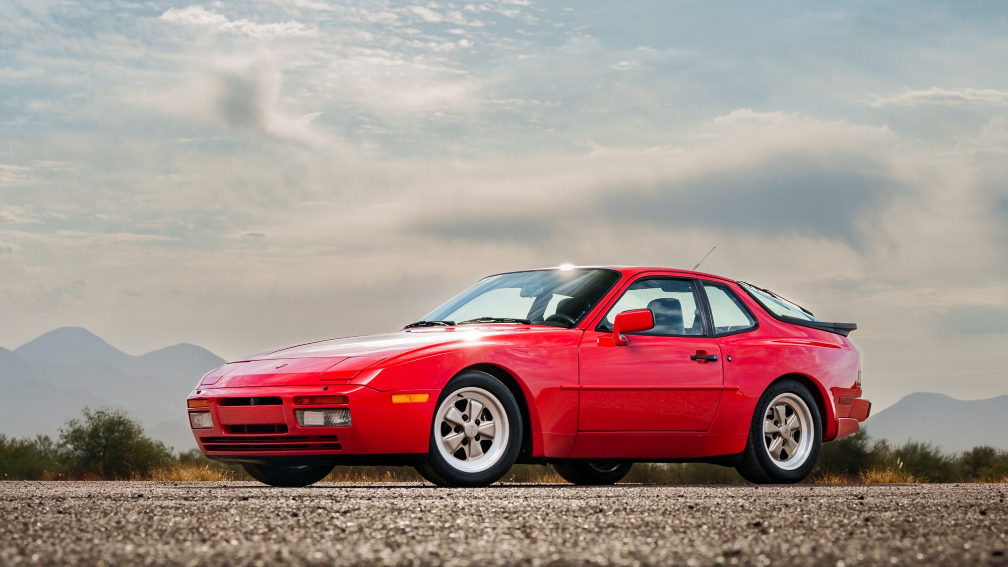 A red Porsche 944 Turbo from the front three-quarter angle on a gravel lot, desert scrub and hazy mountains behind it