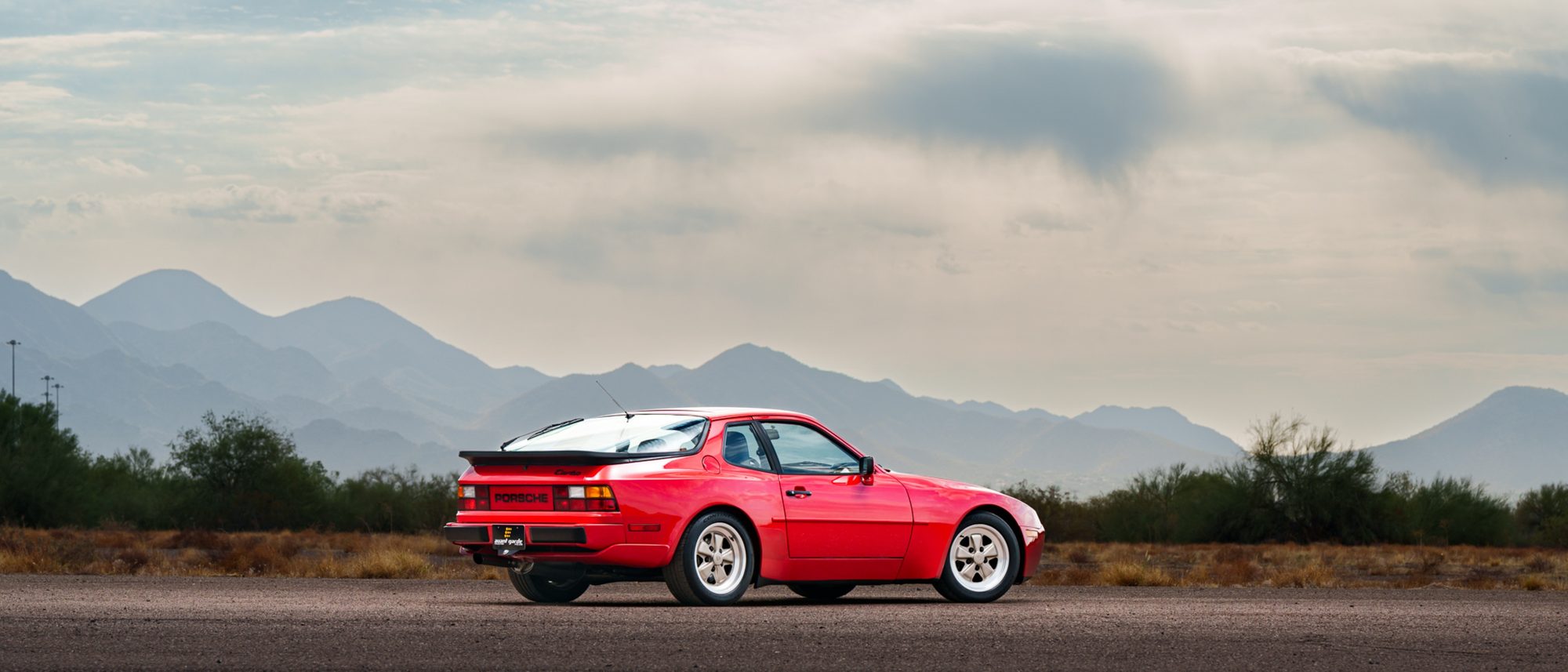 A red Porsche 944 Turbo from the rear three-quarter angle, PORSCHE lettering across its rear light panel, desert scrub and mountains behind it