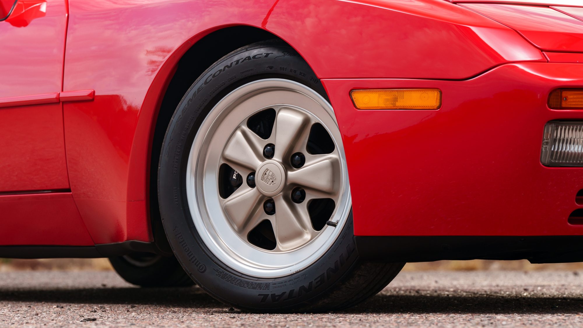 Close-up of a red Porsche 944's front fender, amber side marker and five-spoke alloy wheel with a Porsche crest center cap