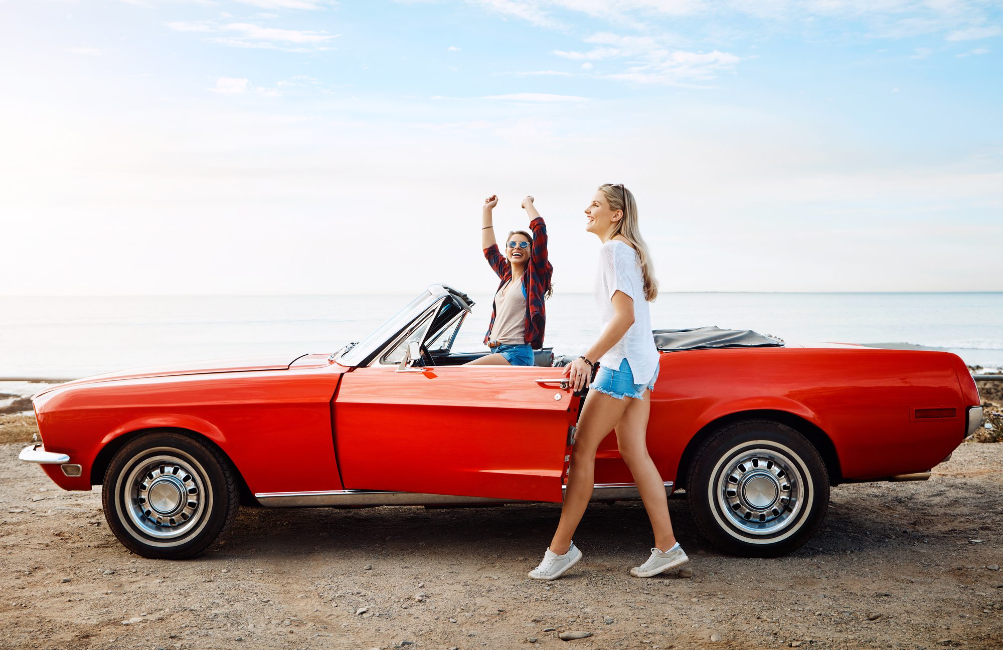 A woman in a white top and denim shorts walking past the open door of a red classic convertible parked on gravel by the sea, while her friend stands in the car with both fists raised