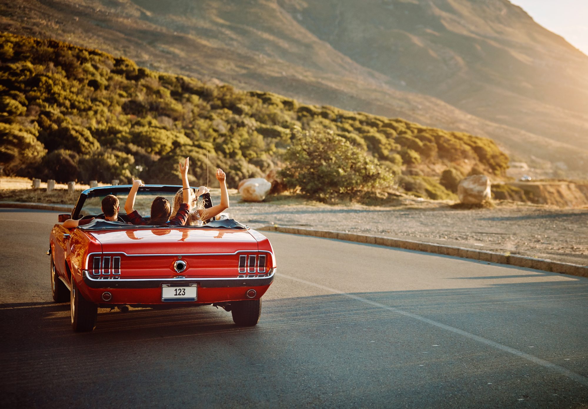 A red 1960s Ford Mustang convertible seen from behind on a coastal road, the driver at the wheel and two passengers with their arms raised