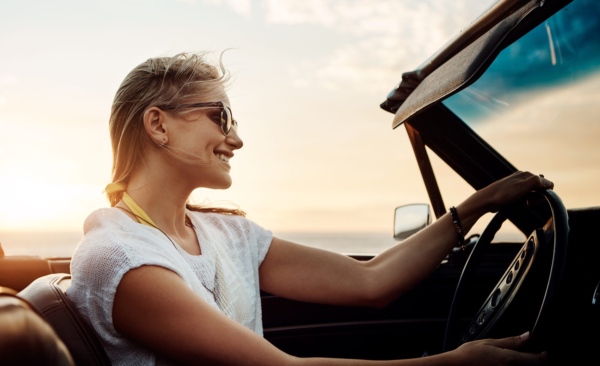 A smiling woman in sunglasses driving an open convertible with both hands on the wheel, the sea on the horizon and a low sun behind her